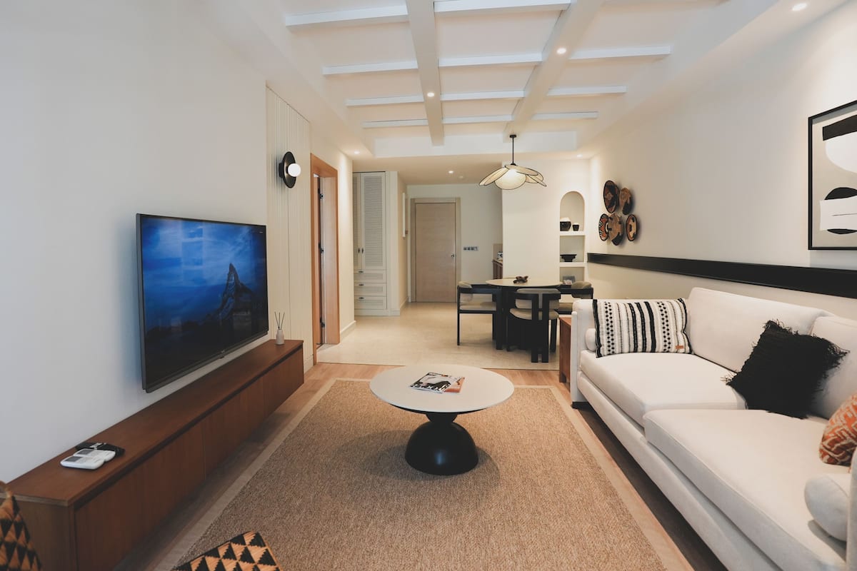 A modern living room with a flat-screen TV, a sofa, a coffee table, and a dining area with a table and chairs.