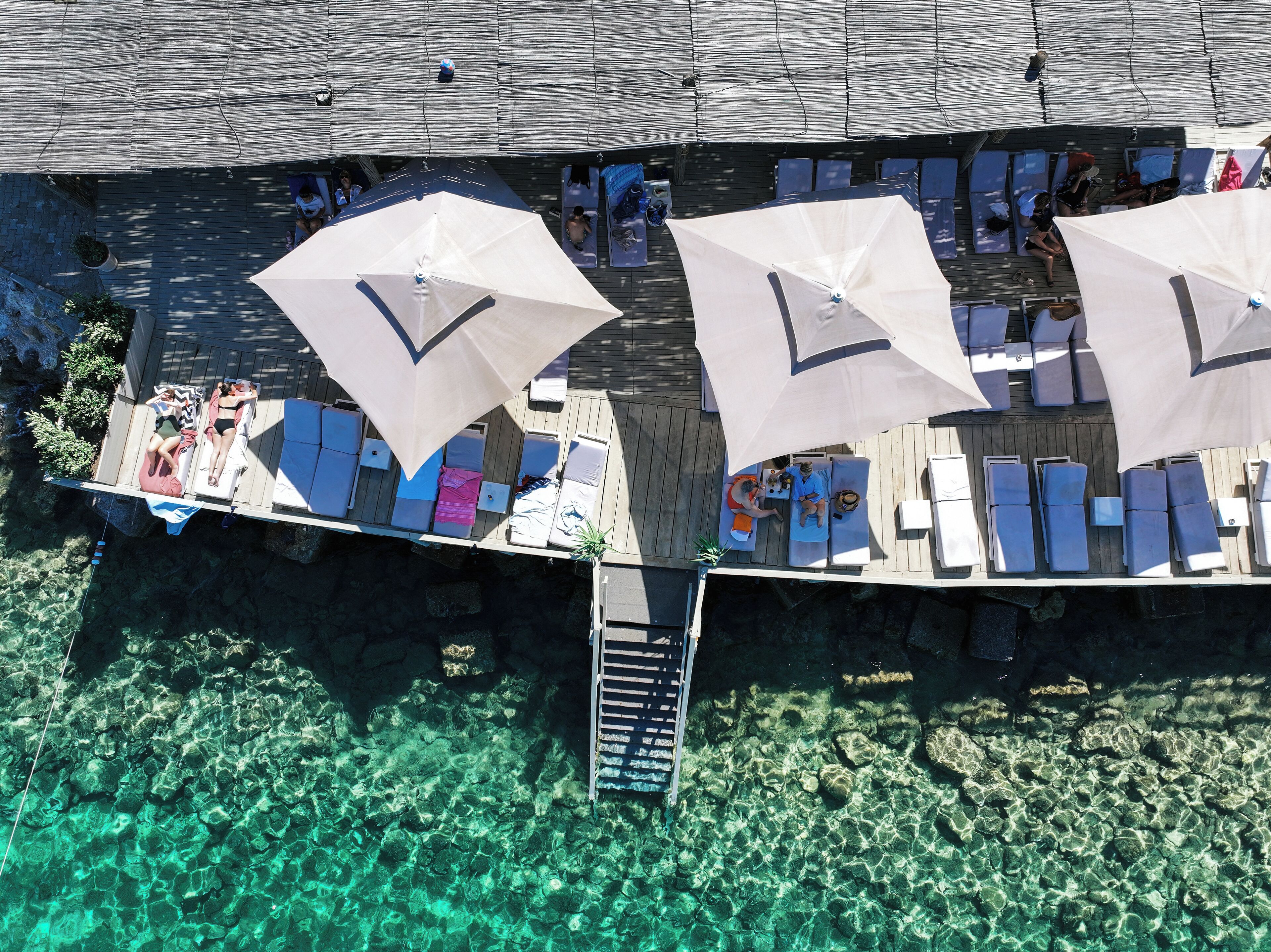 An aerial view of a wooden deck with lounge chairs and umbrellas, overlooking clear turquoise water.
