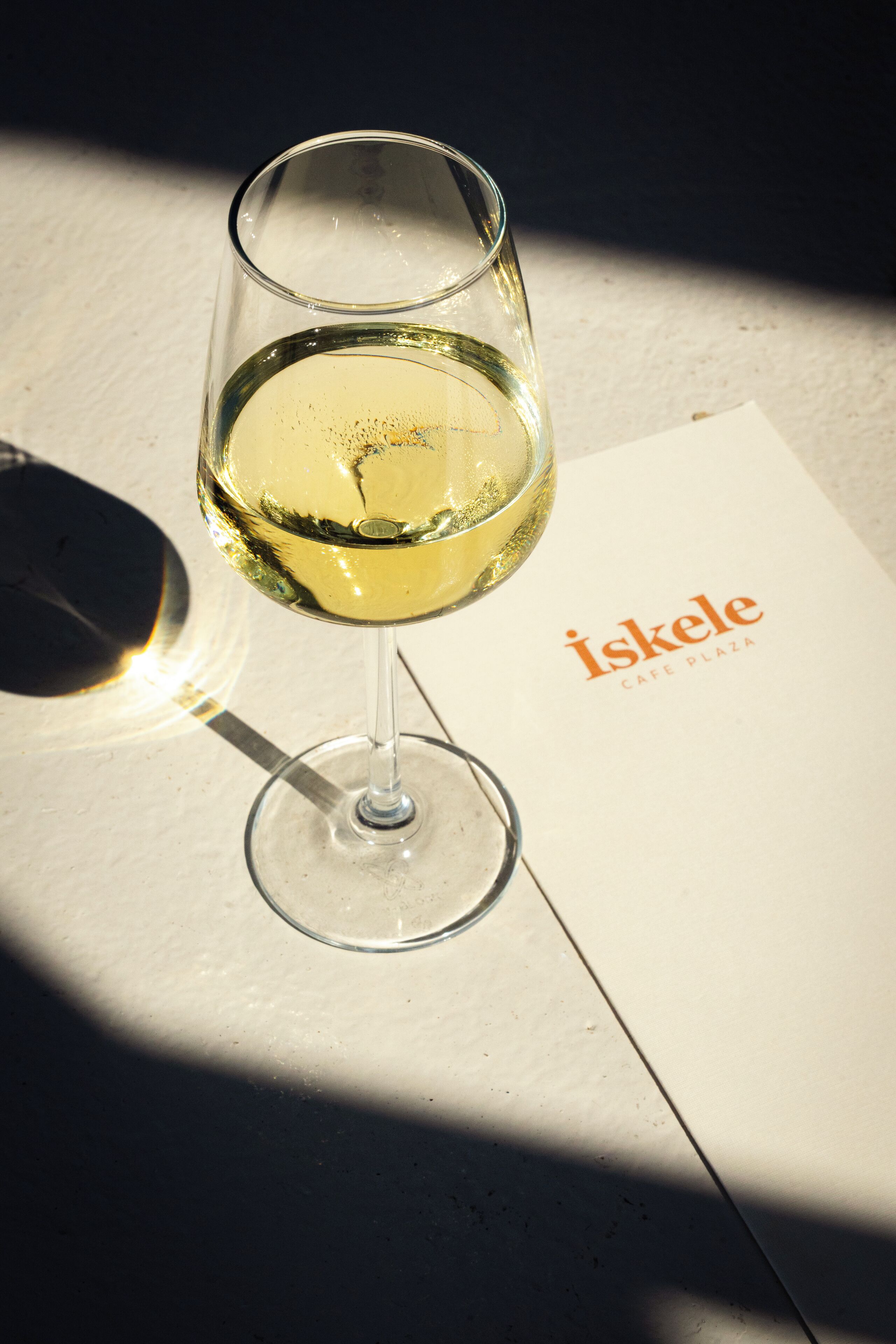 A glass of white wine on a table next to a menu from Iskele Cafe Plaza.
