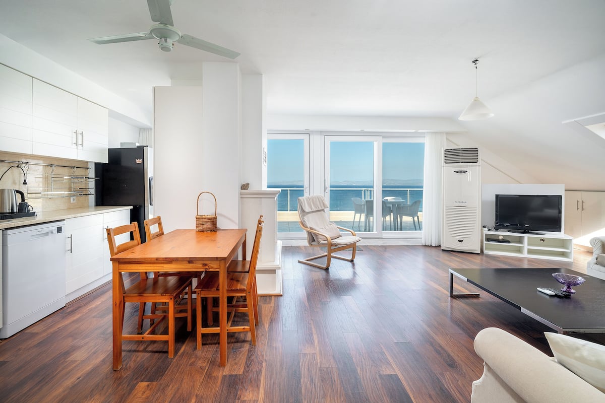 A modern kitchen with white cabinets, a dining table with chairs, a large window with a view of the sea, a living area with a sofa, and a TV.
