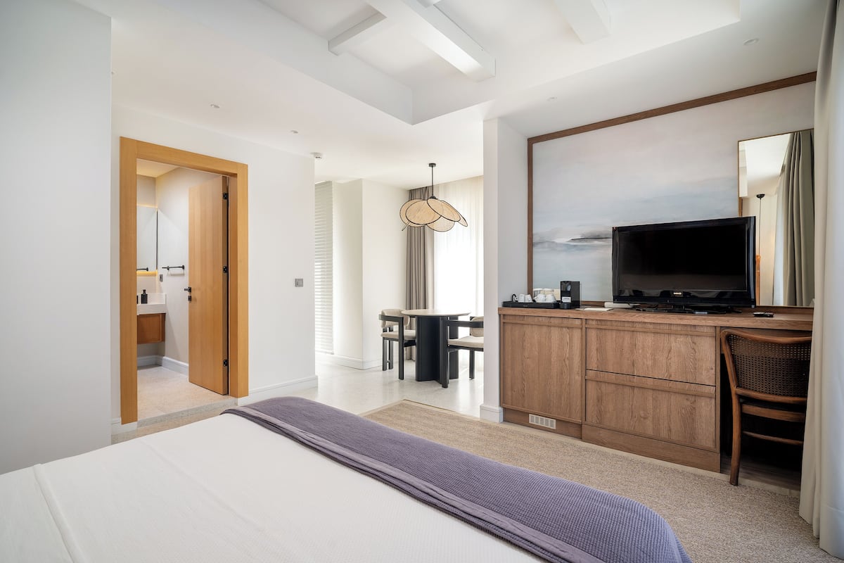A modern hotel room with a large bed, a wooden desk, a television, and a dining area.