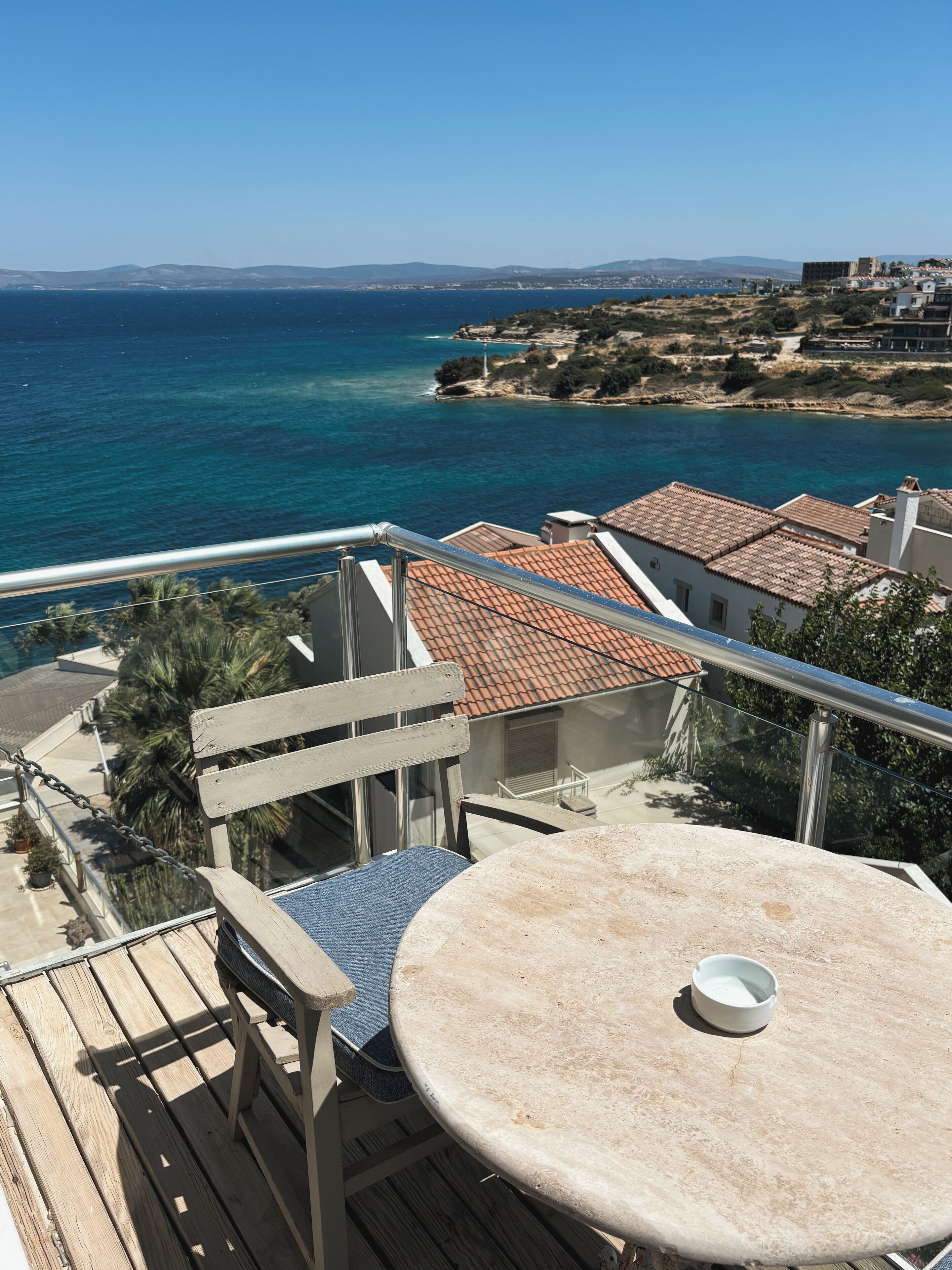 A balcony with a round table and chairs, overlooking a coastal area with buildings and a clear blue sea.