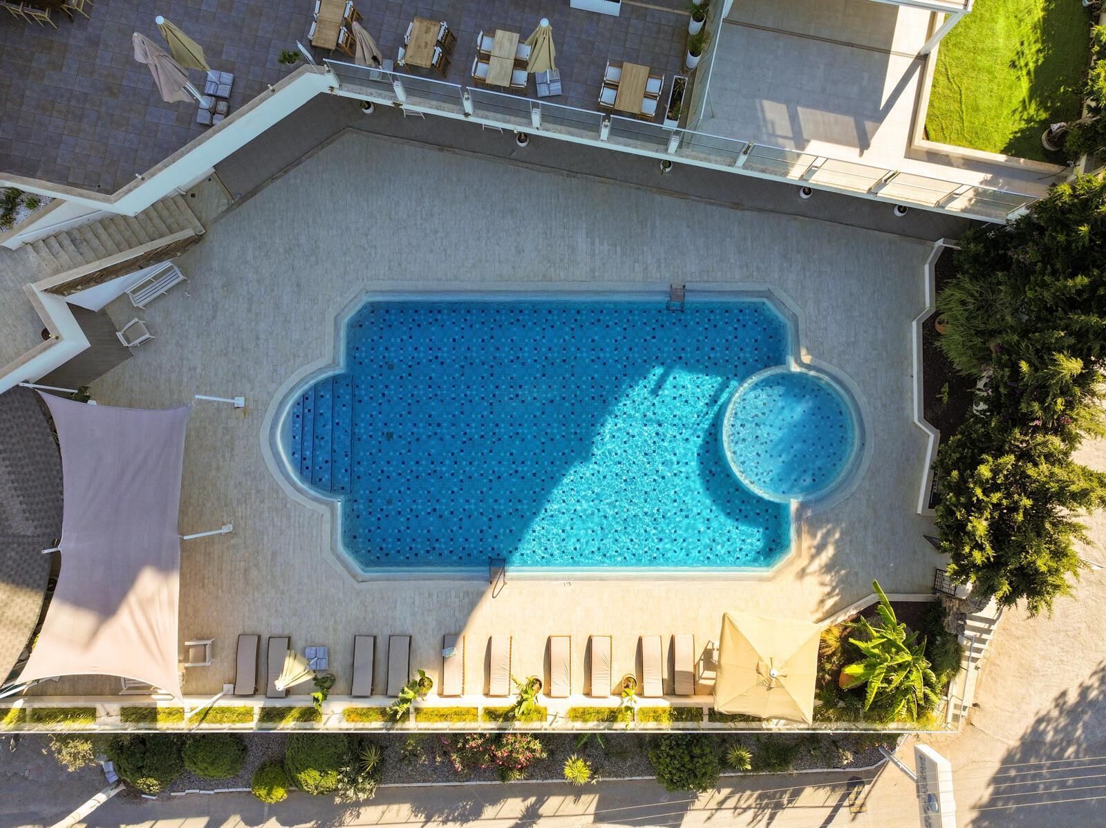 En vy från ovan av en rektangulär swimmingpool med ett nedsänkt bubbelbad, omgiven av solstolar och parasoller.