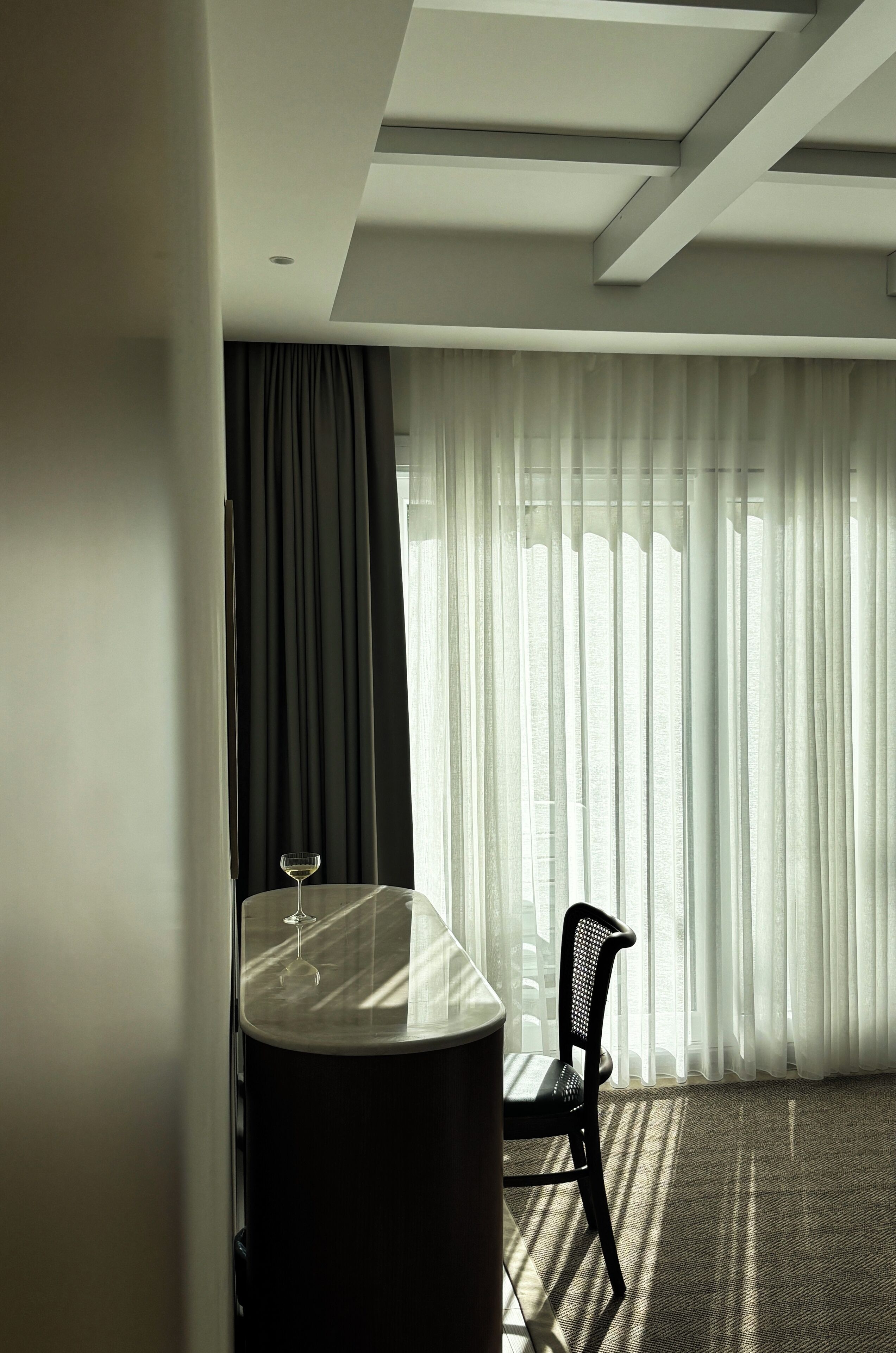 Habitación de hotel con una mesa de comedor, una silla, una copa de vino y un gran ventanal con cortinas traslúcidas.