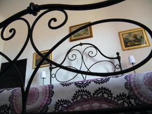 A bed with a metal frame, a patterned bedspread, and two framed pictures on the wall behind. - B&B Il Glicine Residenza D'Epoca (Torrita di Siena)