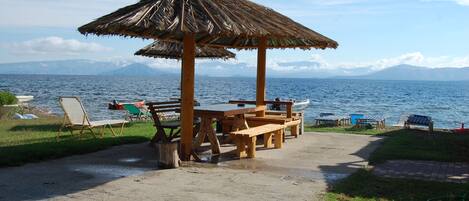 A beachside picnic area with a wooden table and benches, surrounded by lounge chairs and a thatched umbrella, overlooking a calm sea with distant mountains.