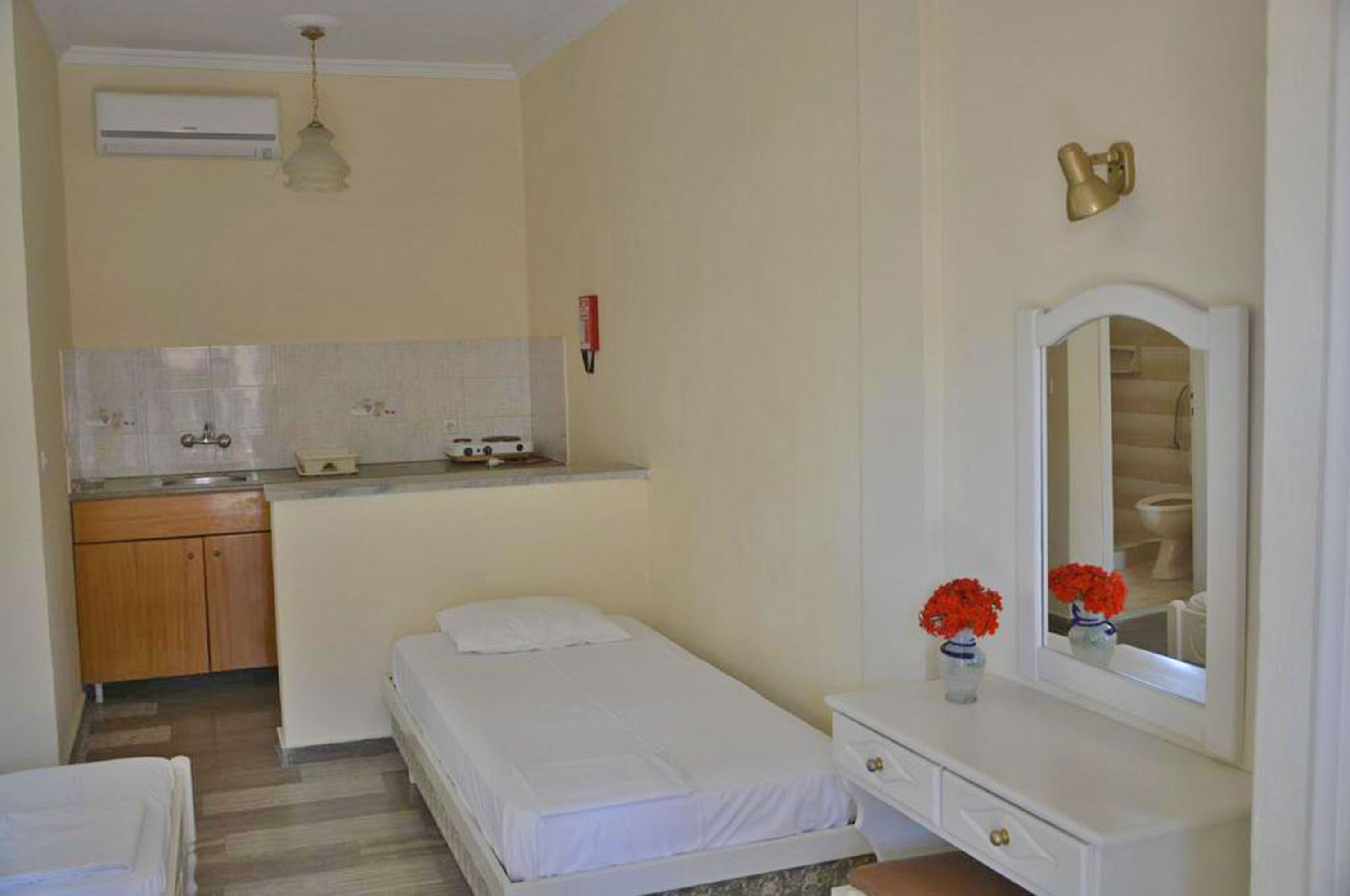 A compact room with a bed, a small kitchenette, a bathroom with a mirror, and a bedside table with flowers.