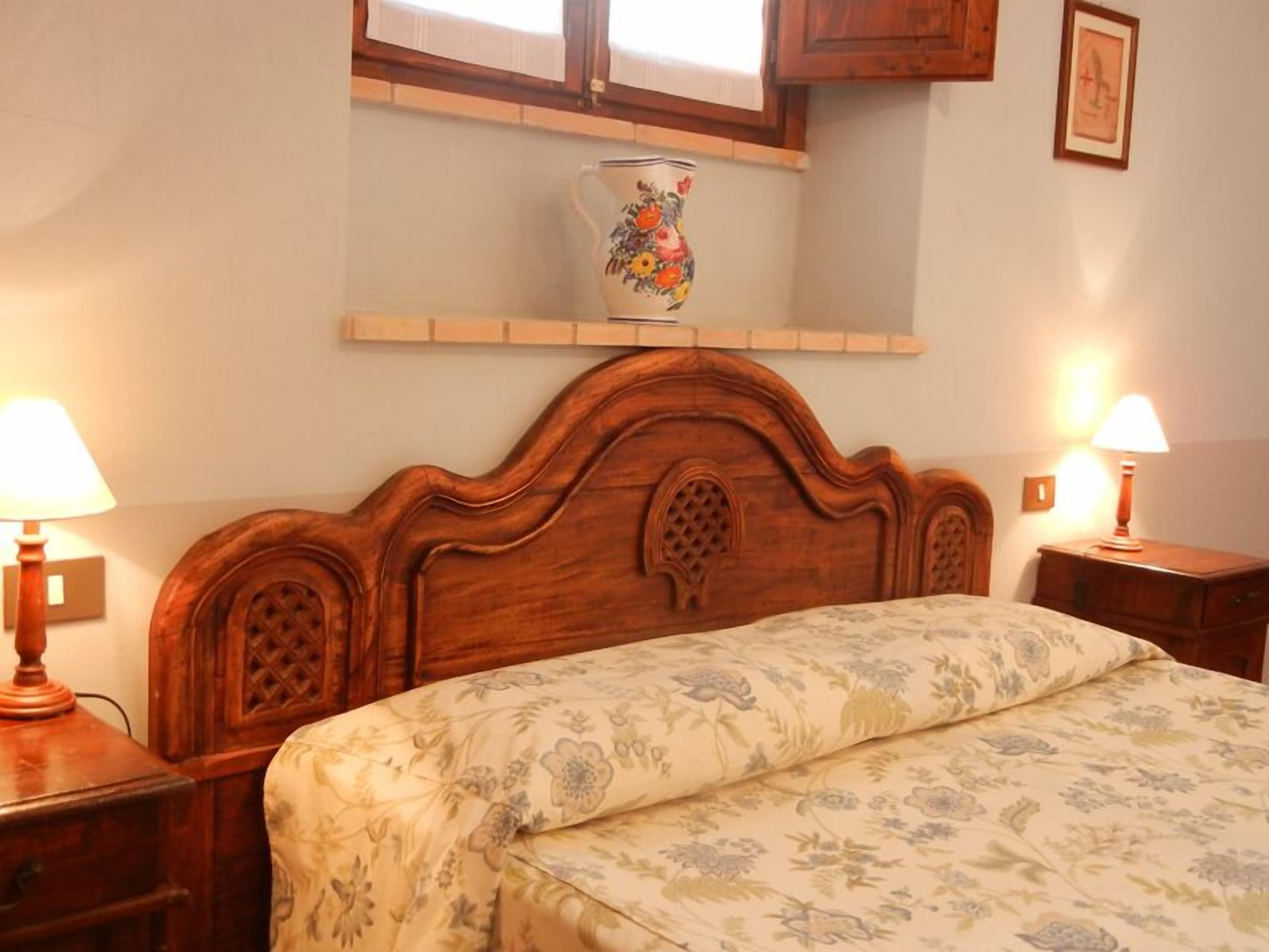 Ein Schlafzimmer mit einem hölzernen Kopfteil, einer blumigen Bettdecke, Nachttischlampen, einem Regal mit einer dekorativen Krug und einem gerahmten Bild an der Wand.