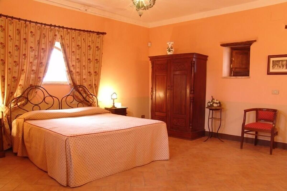 A bedroom with a bed, a wooden wardrobe, a chair, and a small table with a lamp.
