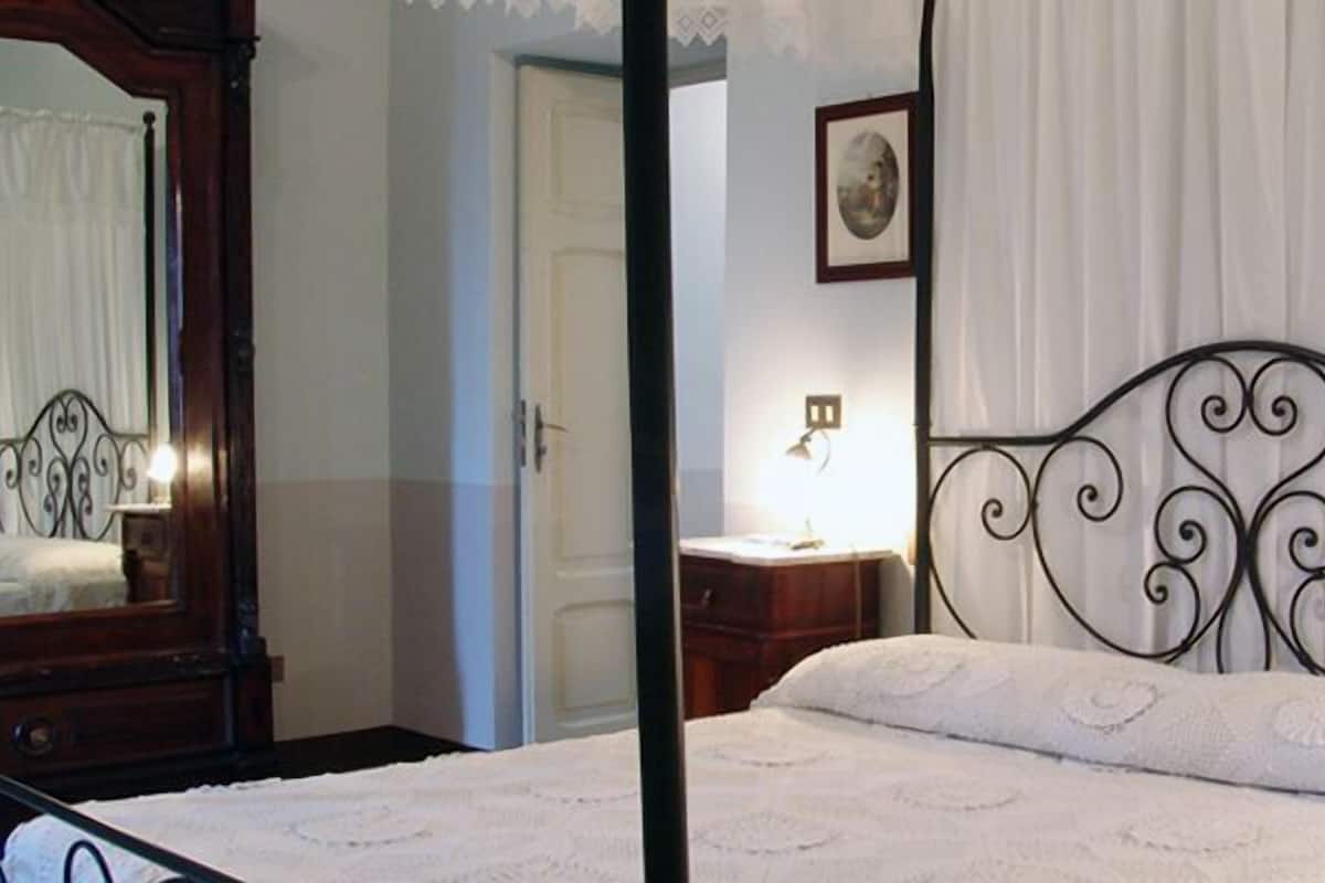 A bedroom with a four-poster bed, a bedside table with a lamp, a mirror, and a framed picture on the wall.