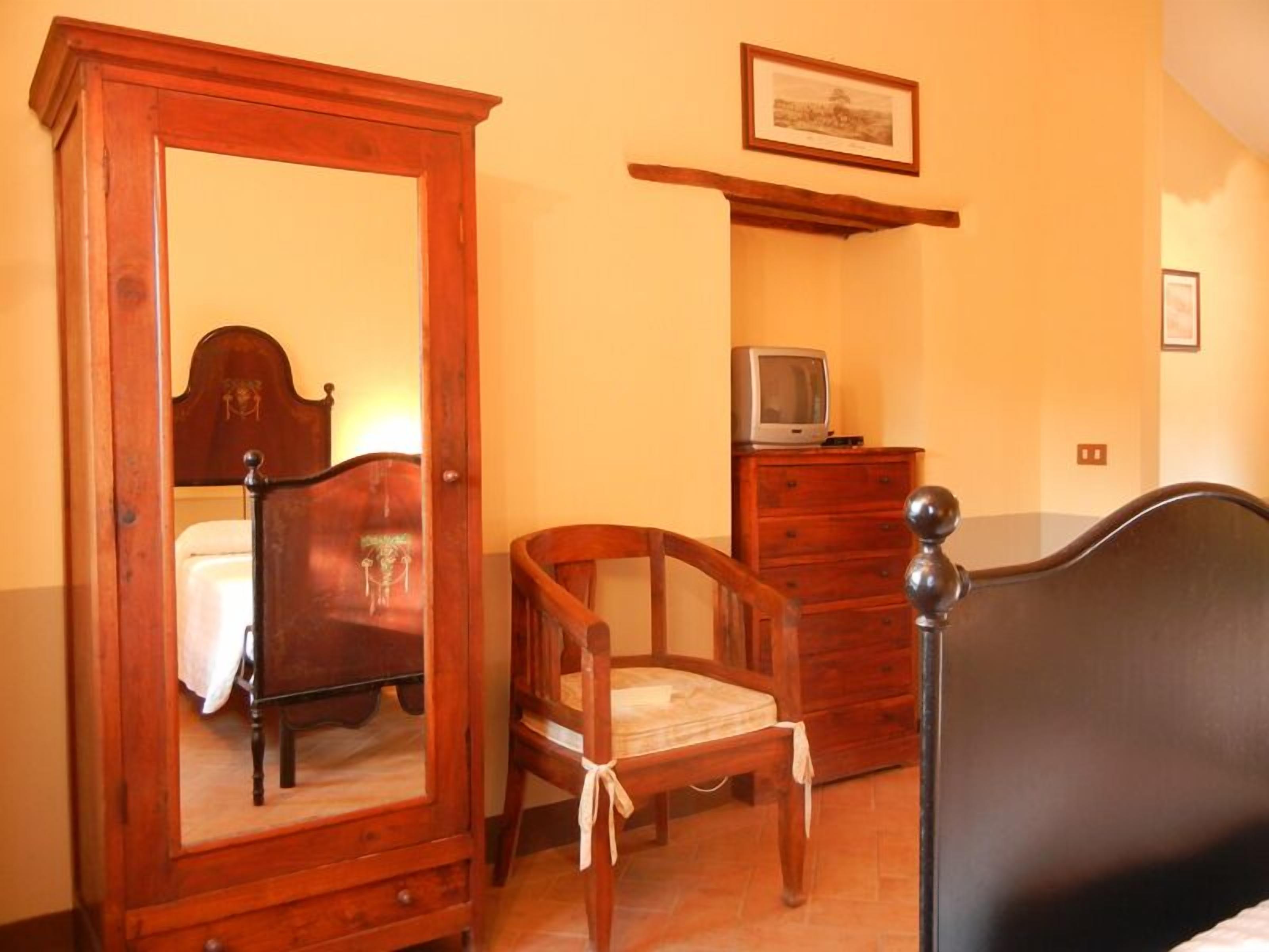 A bedroom with a wooden wardrobe, a bed, a chair, a dresser, and a television on a shelf.