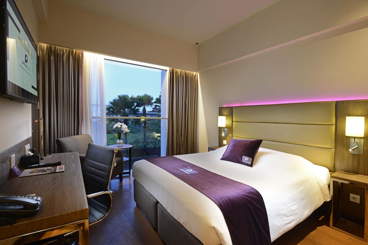 Habitación de hotel con una cama grande, un escritorio con silla, una mesita y vistas a palmeras fuera.