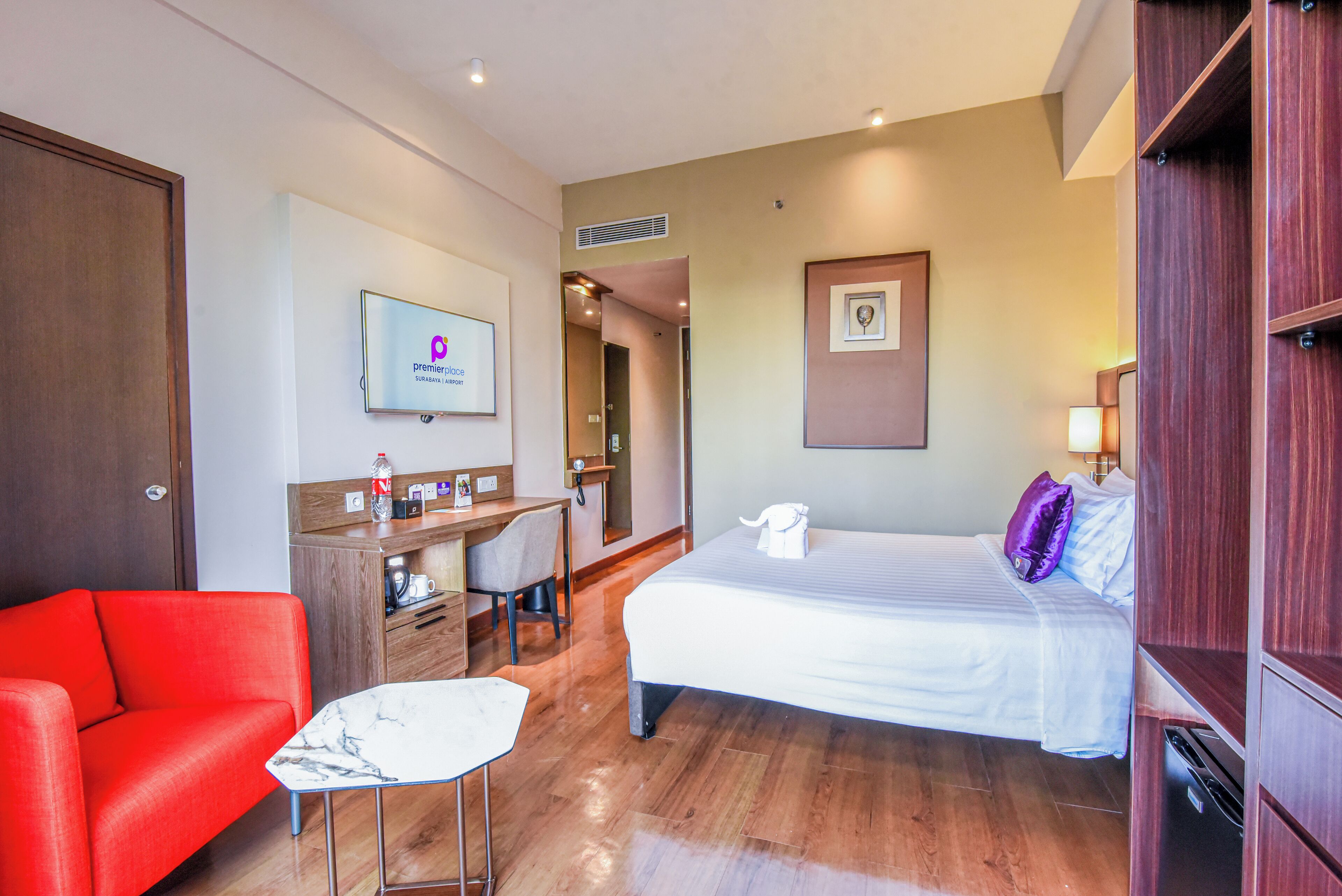 A hotel room with a bed, a desk with a chair, a red sofa, a TV, and a wooden wardrobe.