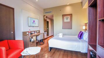 A hotel room with a bed, a desk with a chair, a red sofa, a TV, and a wooden wardrobe.