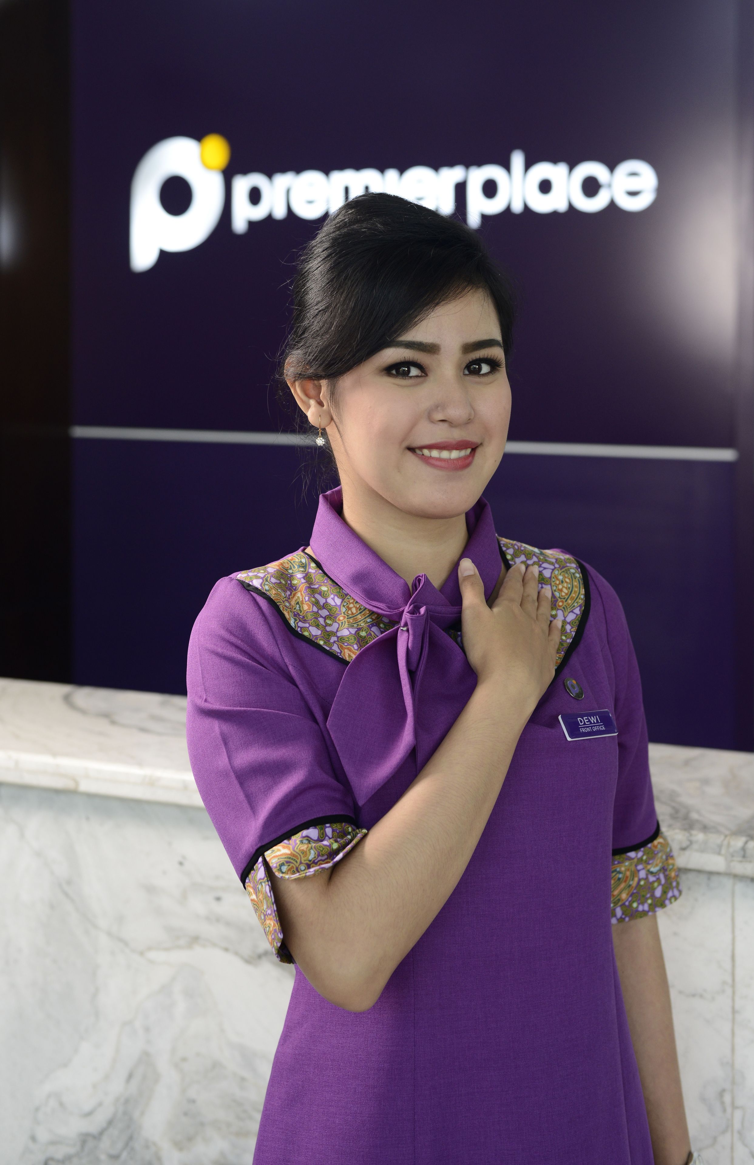 A person in a purple uniform with a name tag, standing in front of a sign with the logo "premerplace".