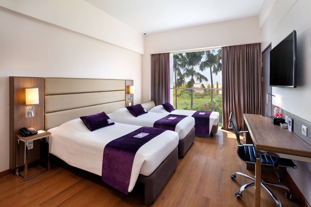 Habitación de hotel con dos camas, un escritorio, un televisor y un balcón con vistas a palmeras.