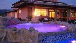 A spa area with a hot tub surrounded by rocks and a building with a dining area visible in the background. - L'Oasi del Fauno Country House (Casal Velino)