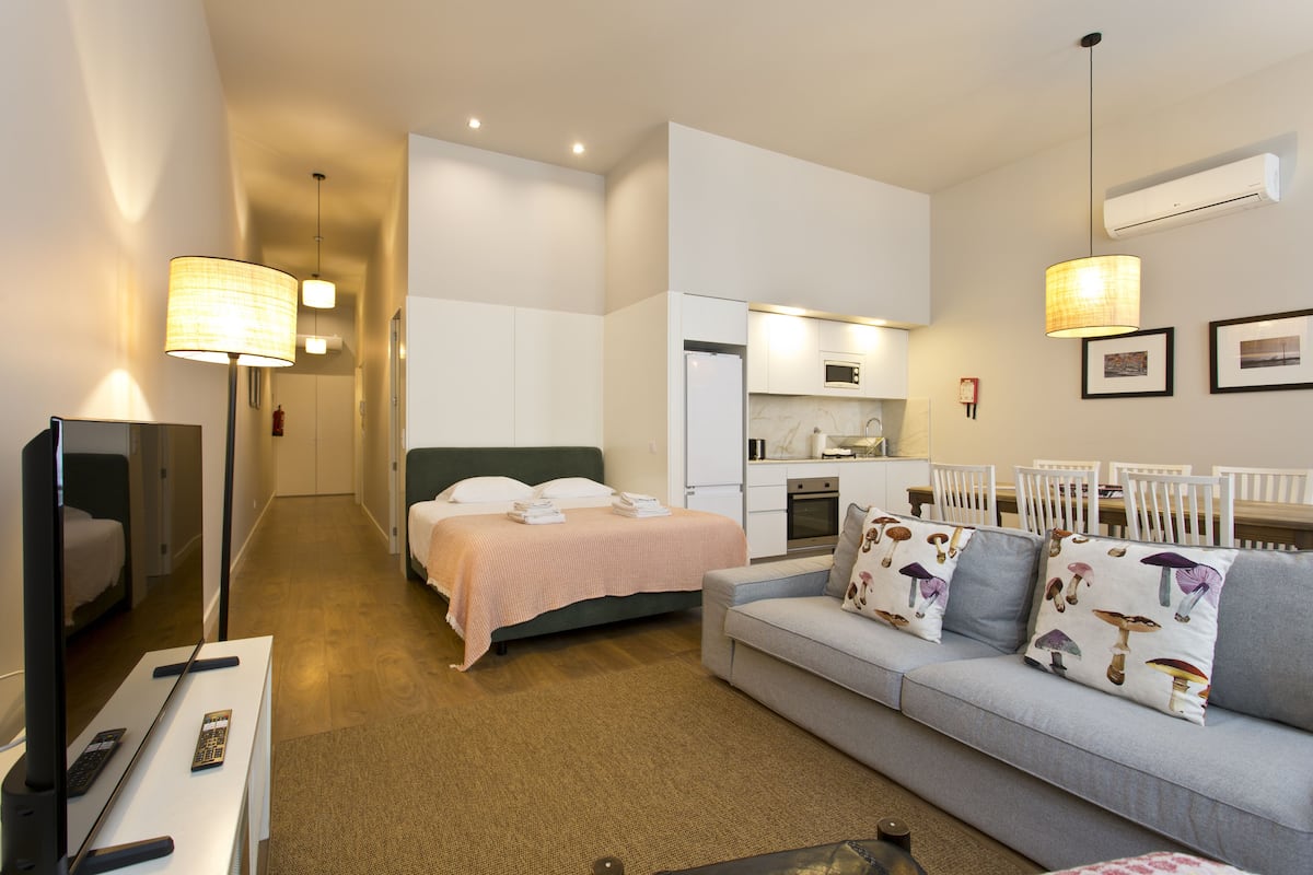 A modern hotel room with a bed, a sofa, a TV, and a kitchenette.
