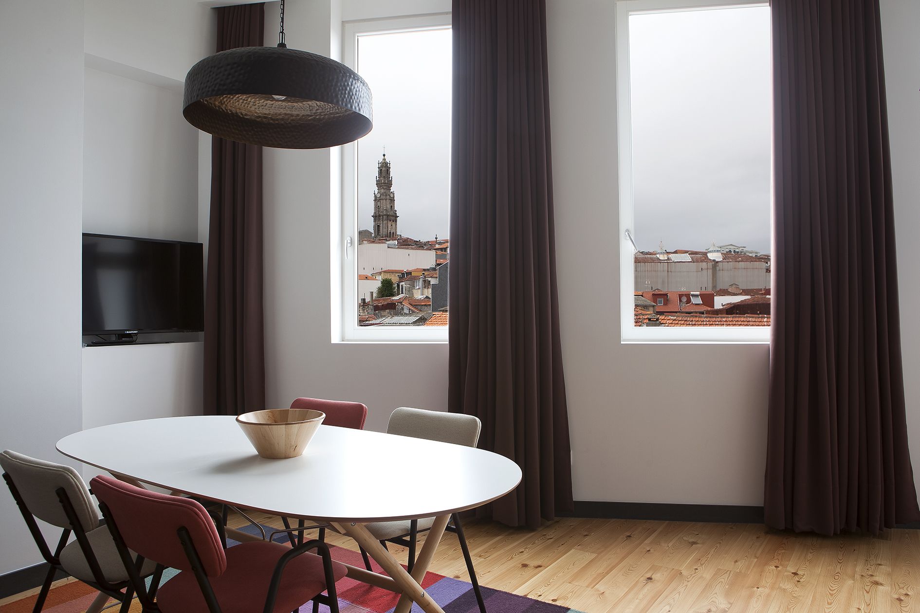 Quarto moderno com uma mesa de jantar redonda, televisão e grandes janelas com cortinas.
