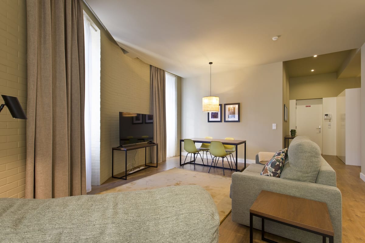 A modern hotel room with a sofa, a small table, a desk with a chair, a TV, and a dining area with chairs.