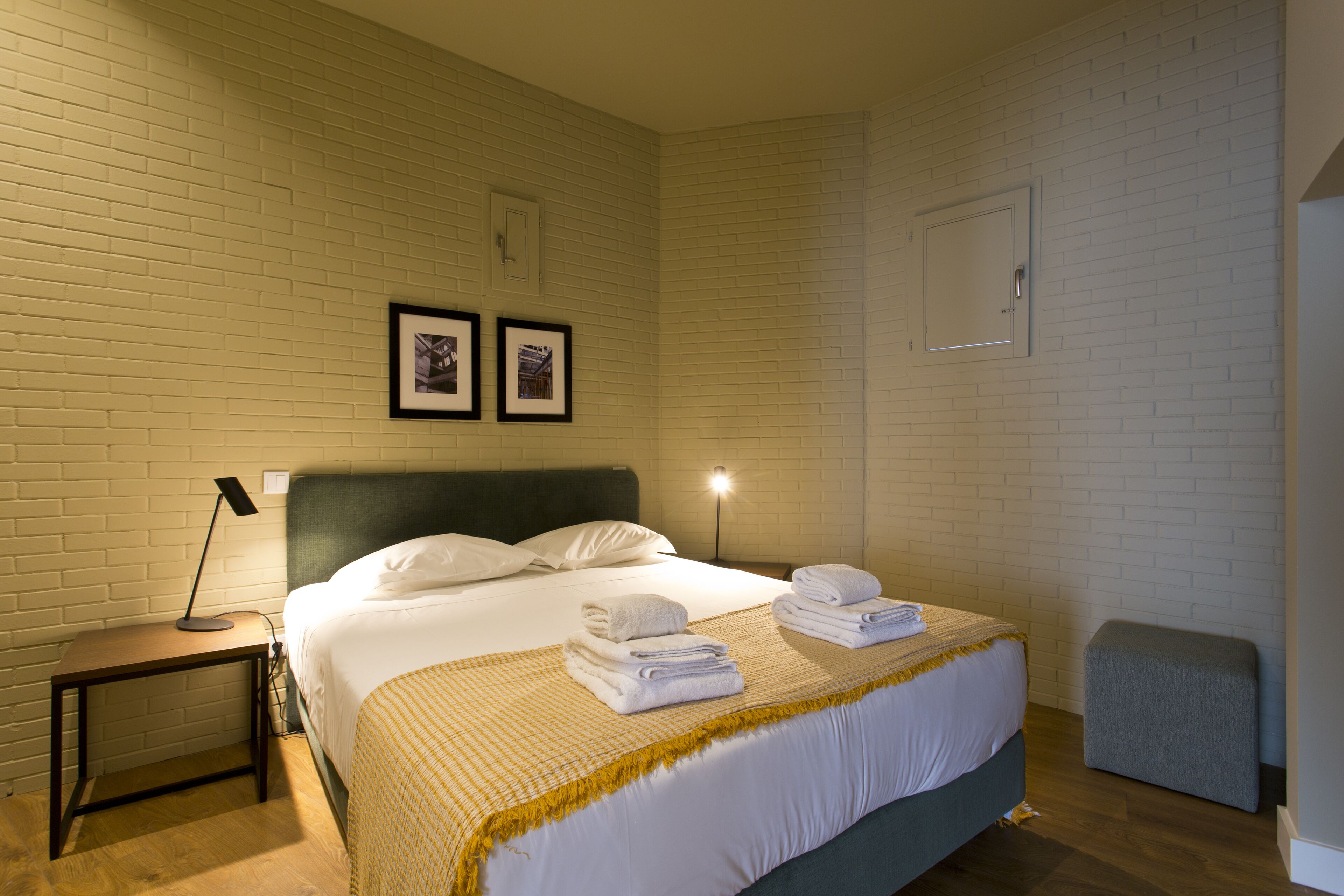 A hotel room with a bed, bedside table, lamp, and a small stool.