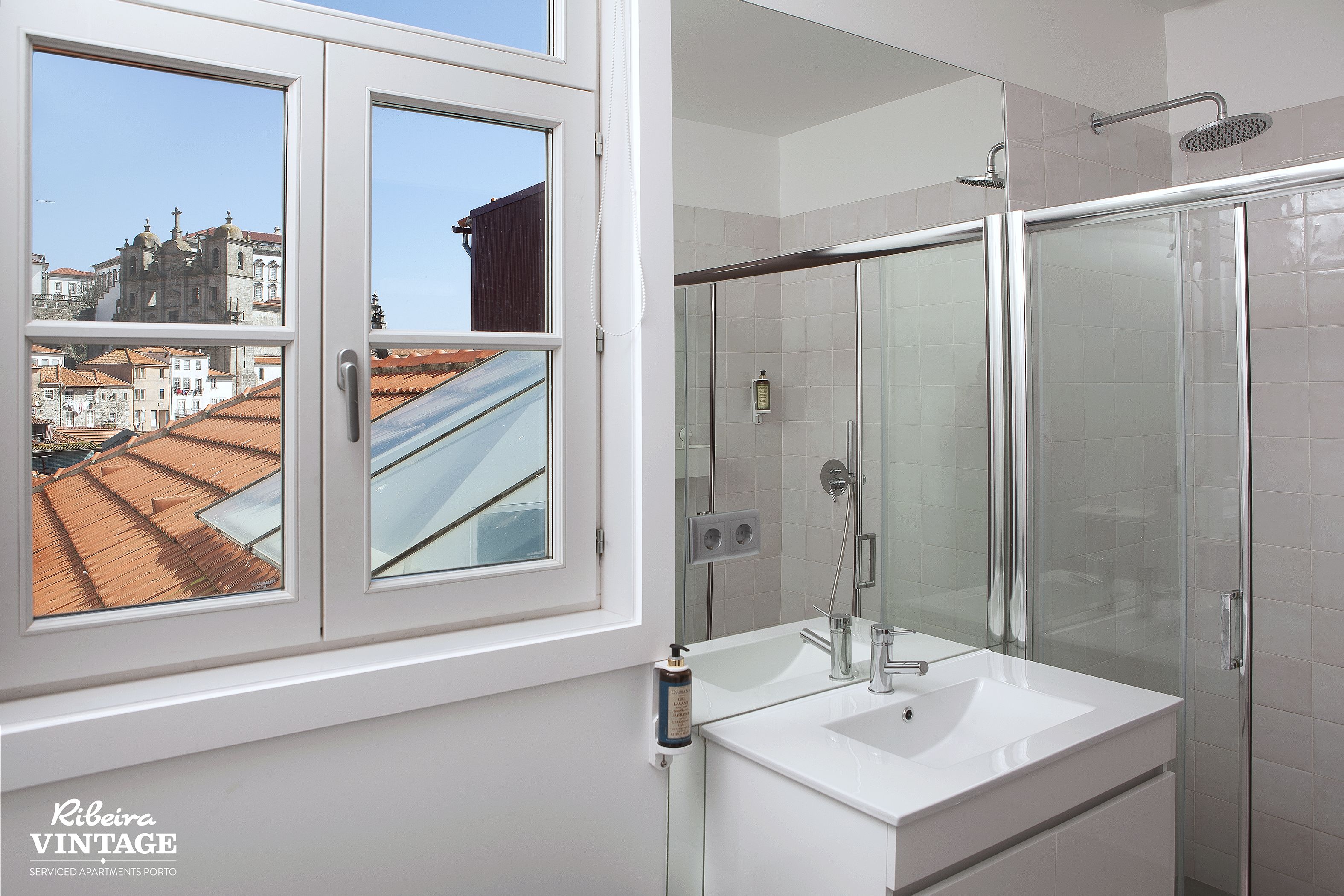 Un baño con un amplio ventanal, cabina de ducha con mampara de vidrio y lavamanos blanco.