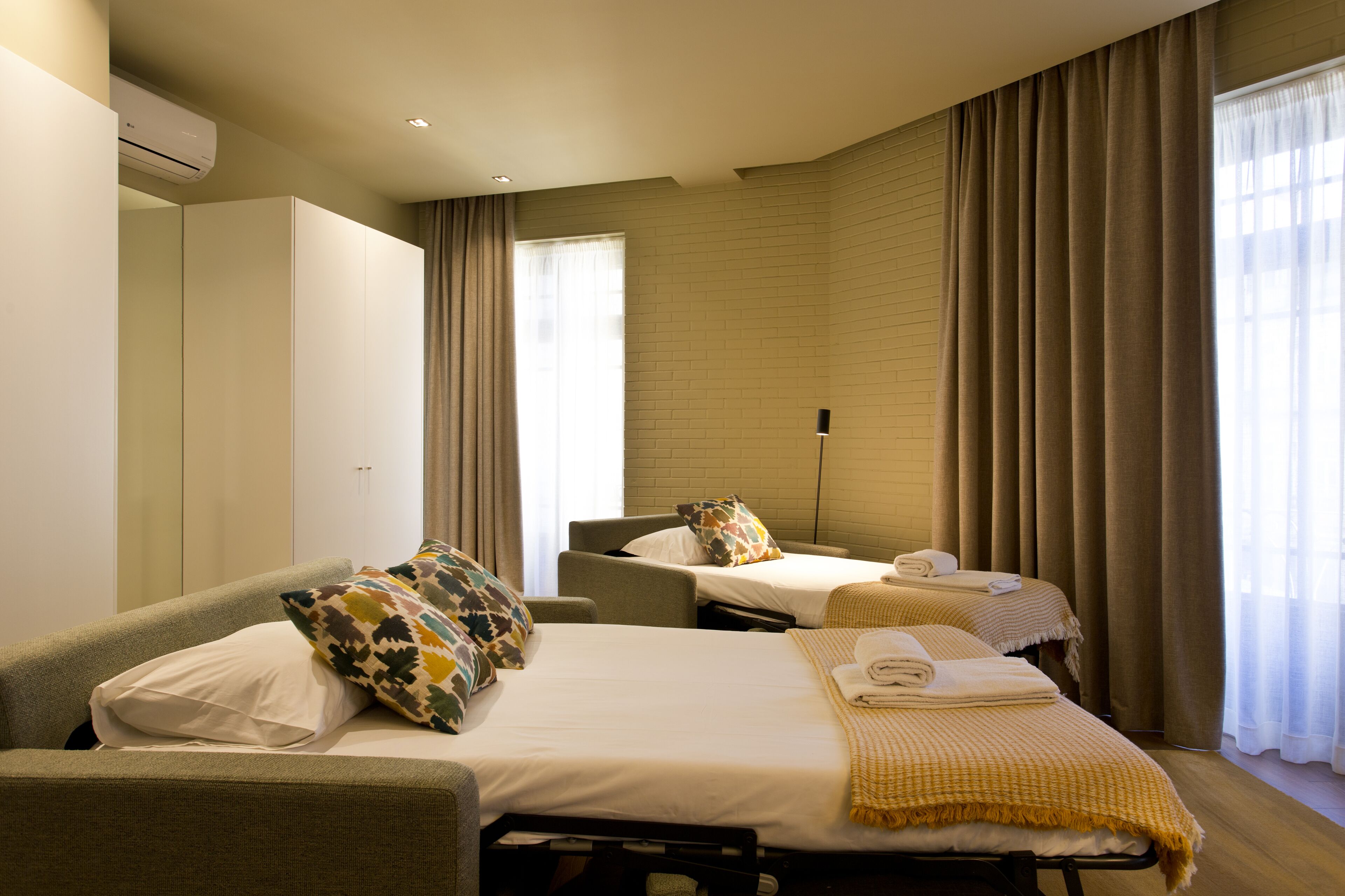 A hotel room with two beds, a sofa, a flat-screen TV, and a large window with curtains.