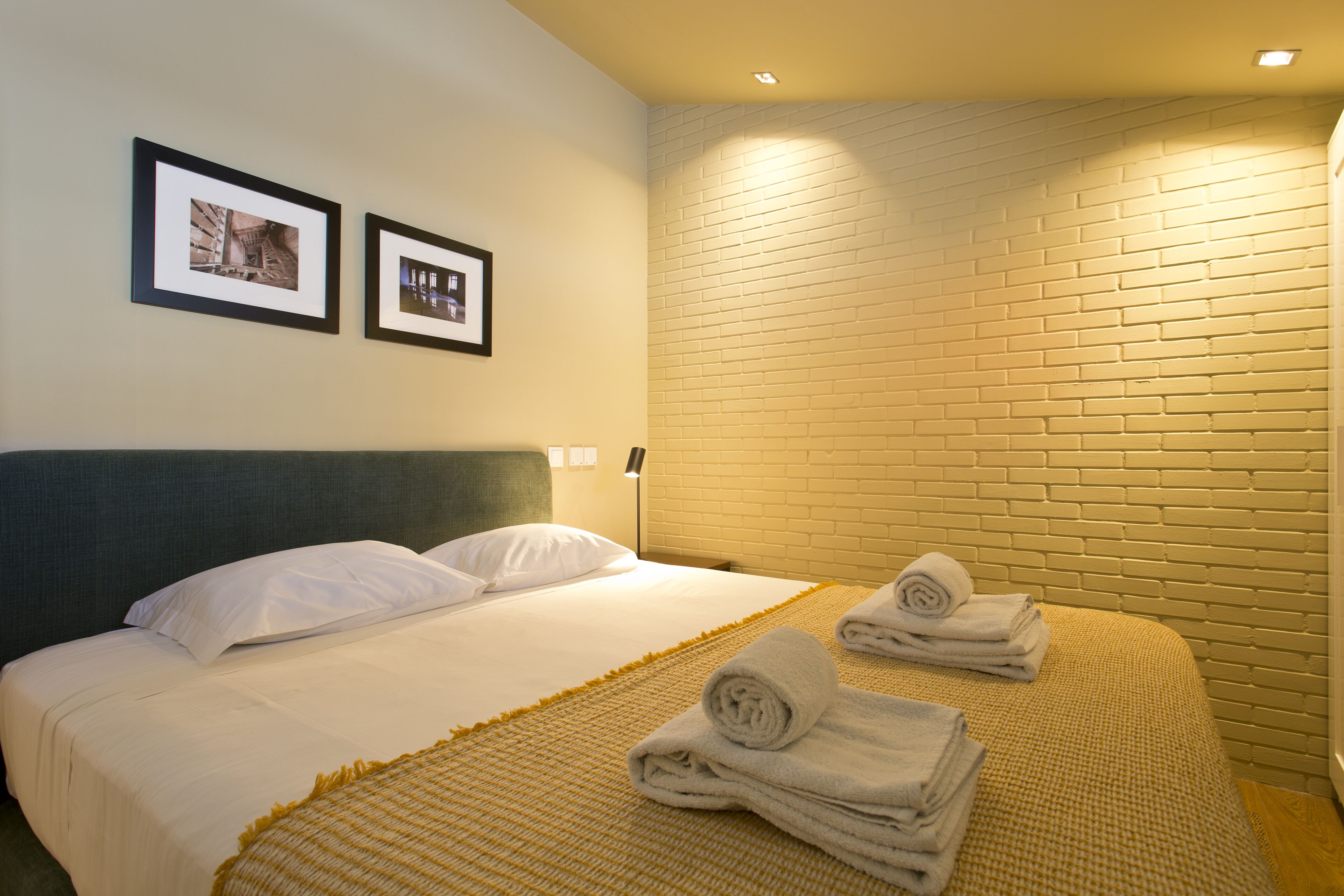 Un lit bien fait, avec du linge de lit blanc et une serviette pliée posée sur le côté, une tête de lit et un mur décoré d’un motif en briques.