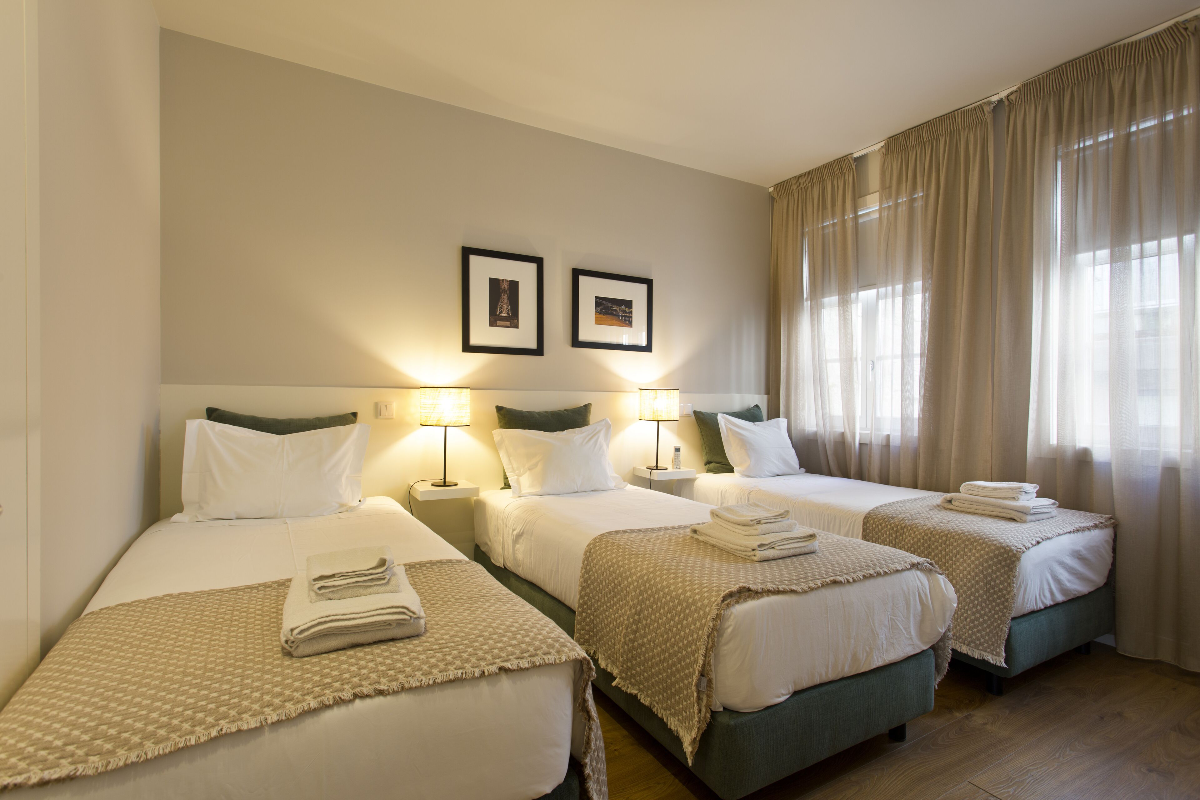 A hotel room with two single beds, a large window with curtains, and two framed pictures on the wall.