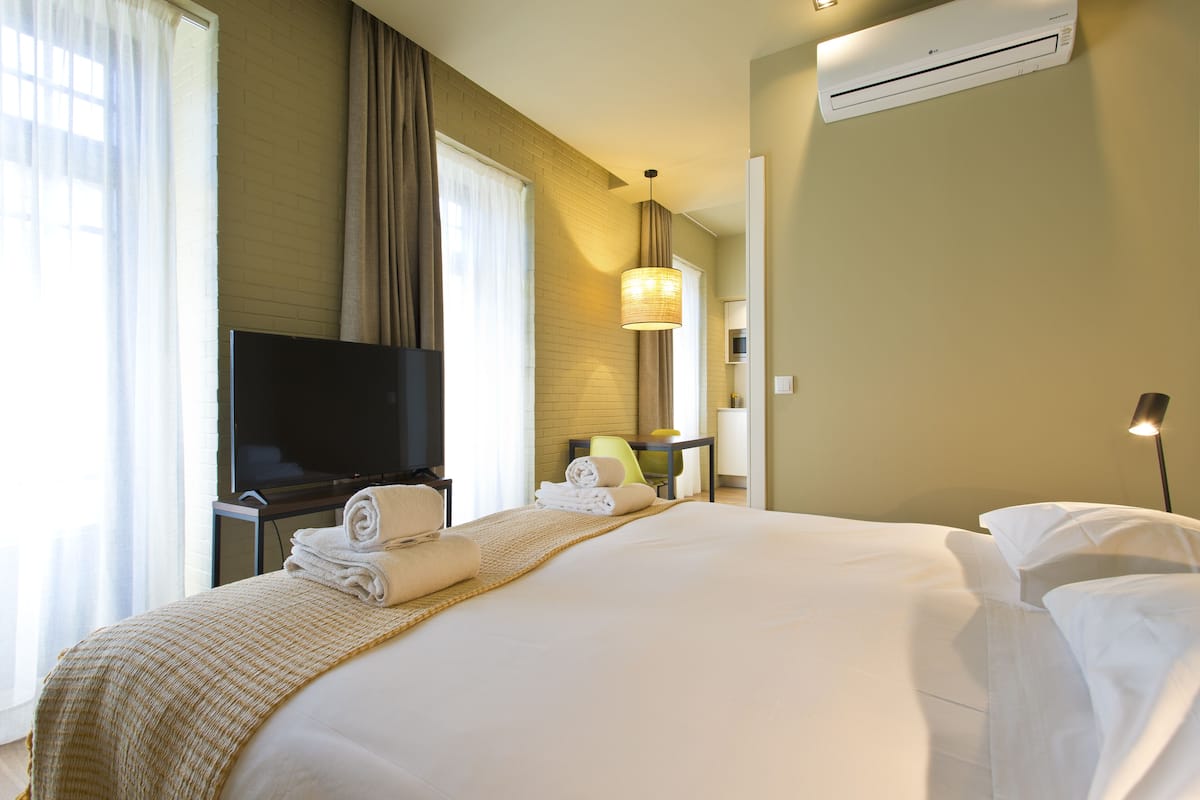 A hotel room with a large bed, a flat-screen TV, a desk with a chair, a lamp, and an air conditioning unit.