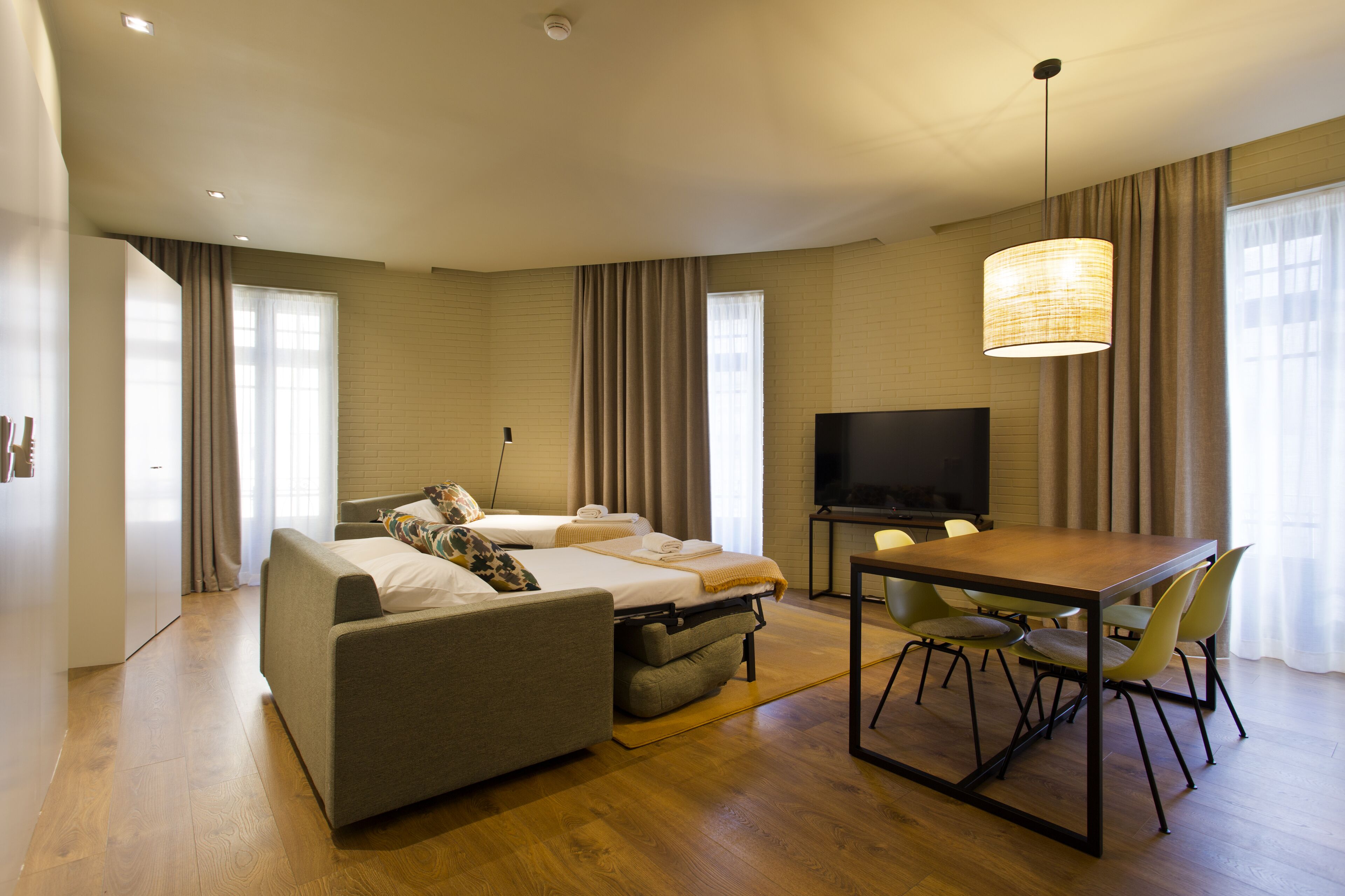A modern hotel room with a sofa, two single beds, a dining table with chairs, a TV, and large windows with curtains.
