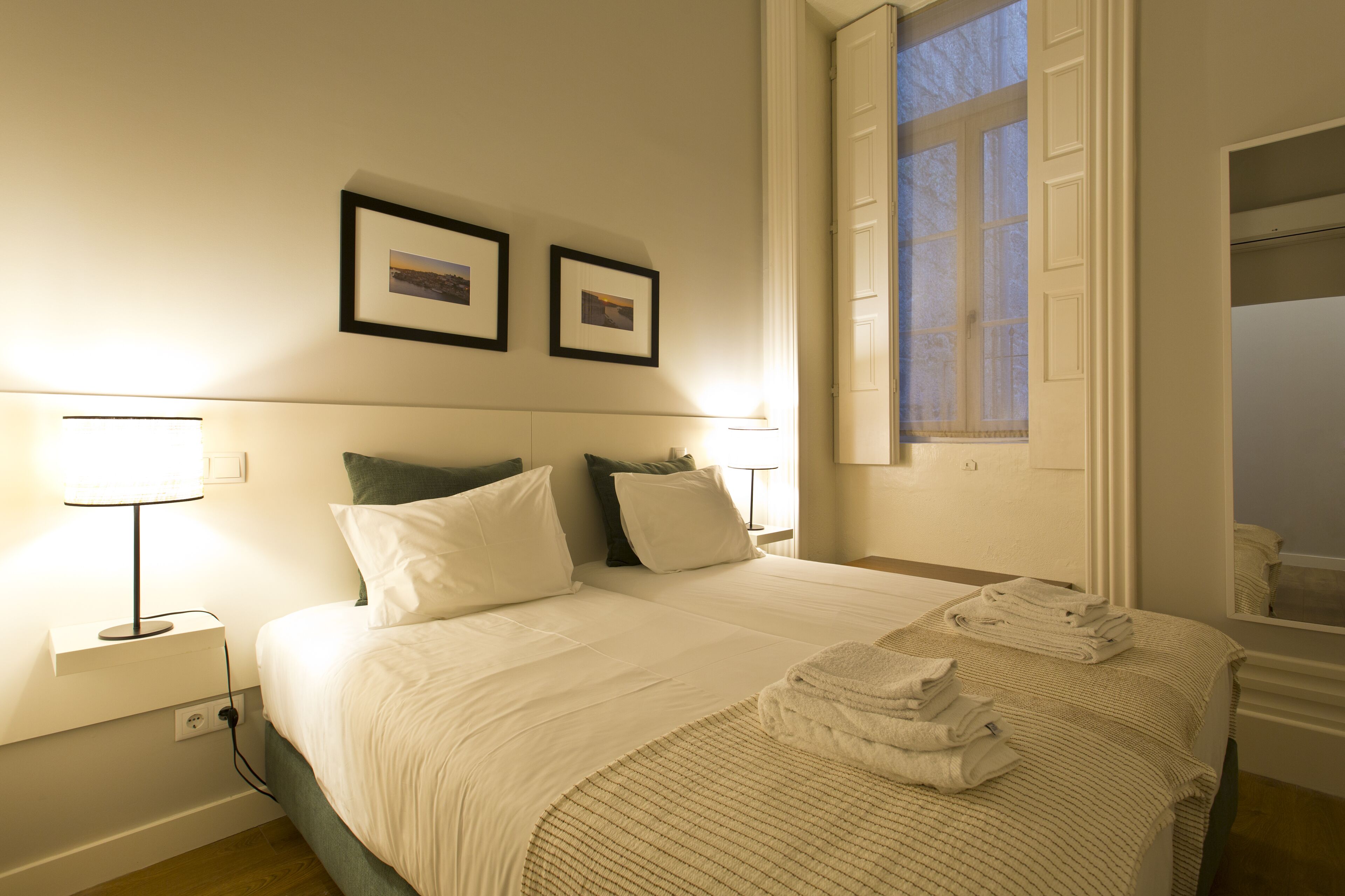 A neatly made bed with white linens and a folded towel on the side. There are two bedside lamps and a window with a view of the outside.