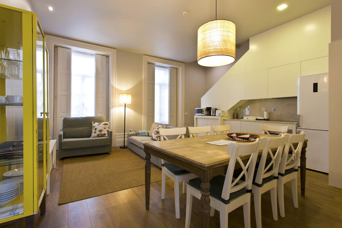 A modern kitchen with white cabinets, a dining table, and chairs. There is a sofa and a lamp in the living area.