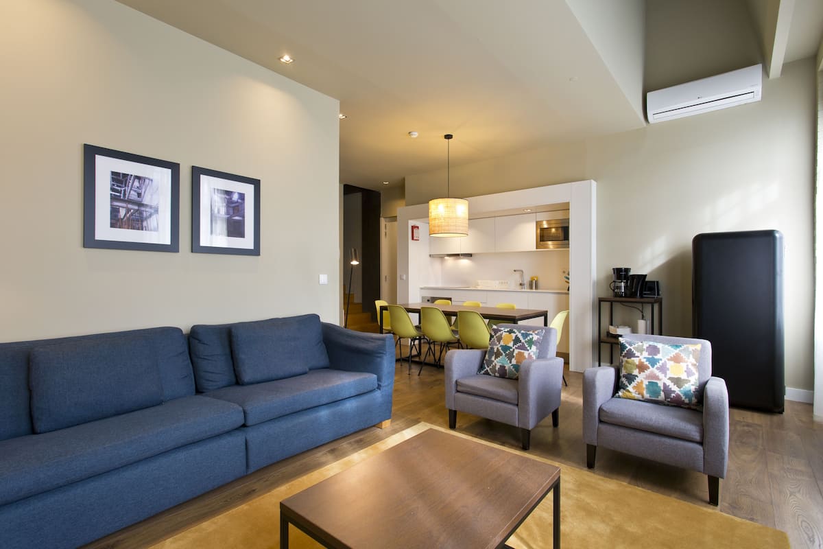A modern living room with a blue sofa, two armchairs, and a coffee table. A kitchen area is visible in the background with a dining table and chairs.