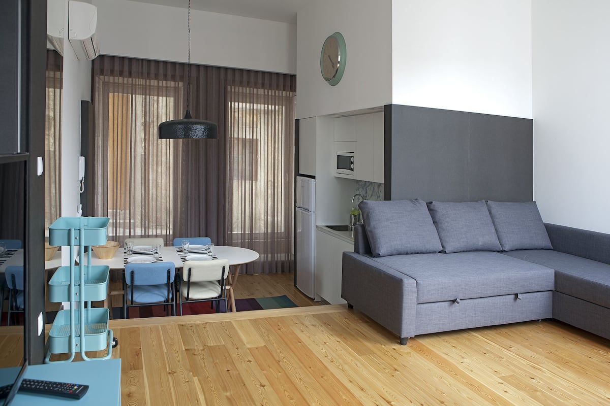 A modern living room with a grey sofa, wooden flooring, a dining area with a table and chairs, and a kitchen area with a microwave and refrigerator.