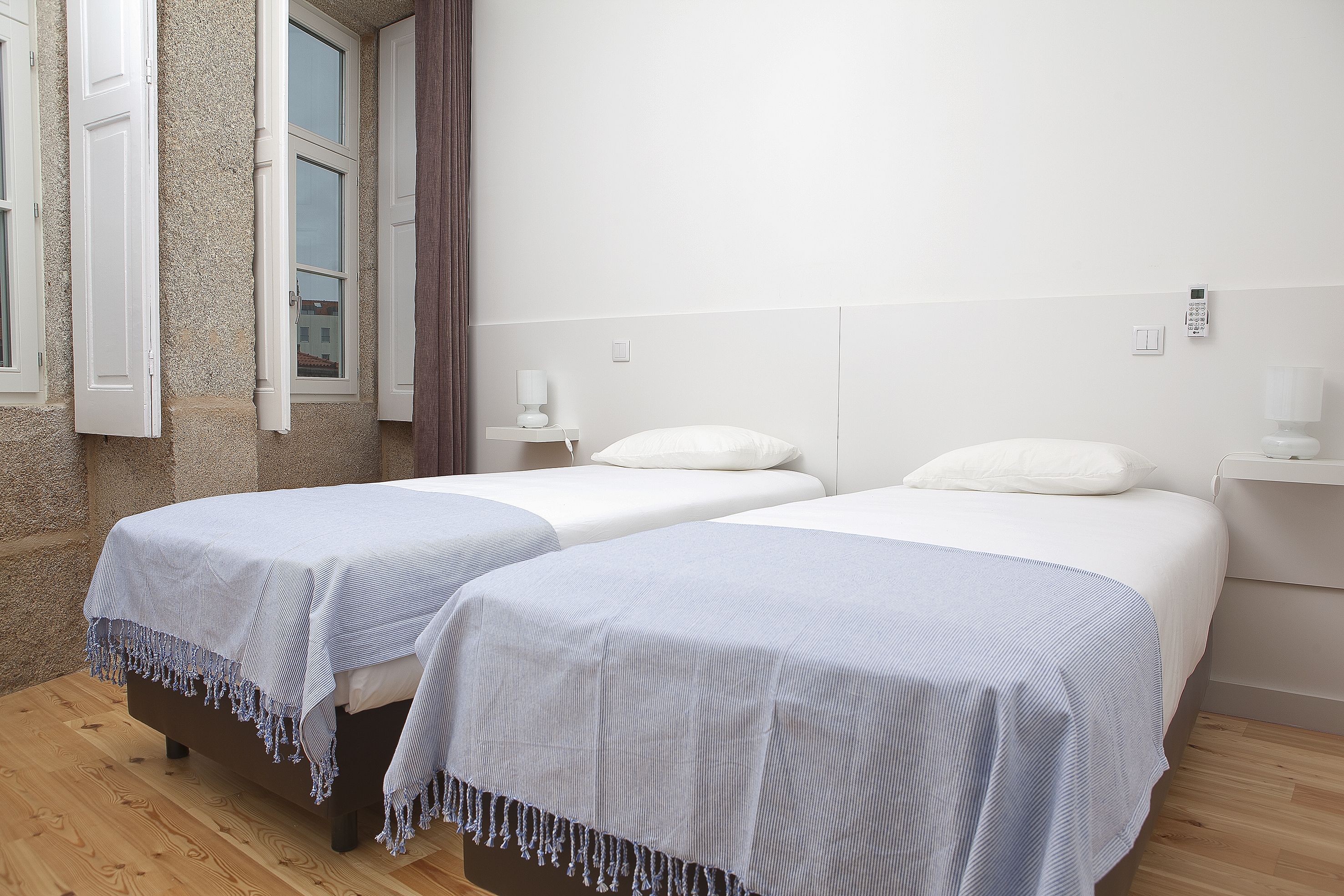 A hotel room with two beds, each with a white headboard and a blue-striped bedspread. There are white nightstands with lamps, and the room has a stone wall and wooden flooring.