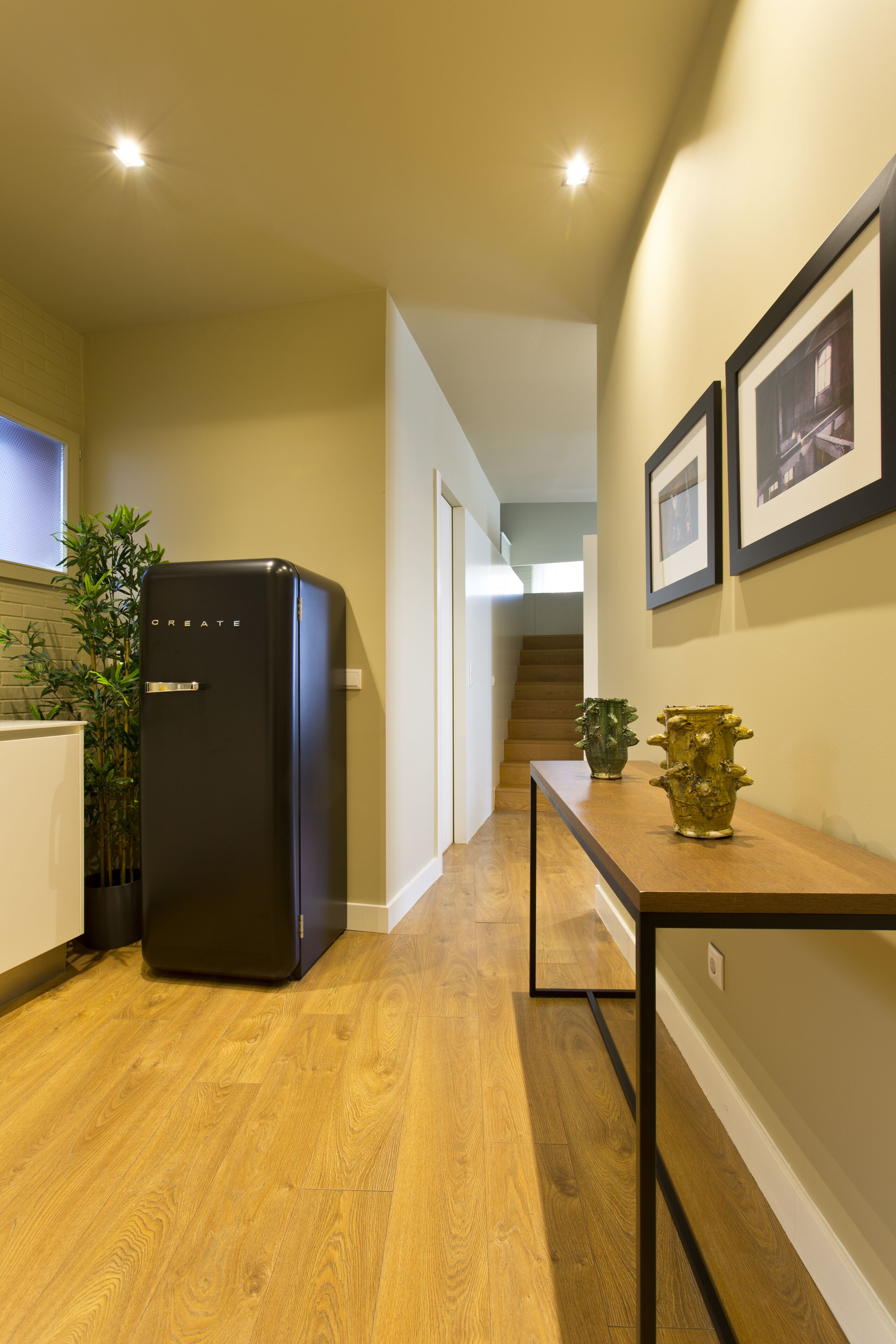 A hallway with a black refrigerator, wooden flooring, a staircase, and framed pictures on the wall.