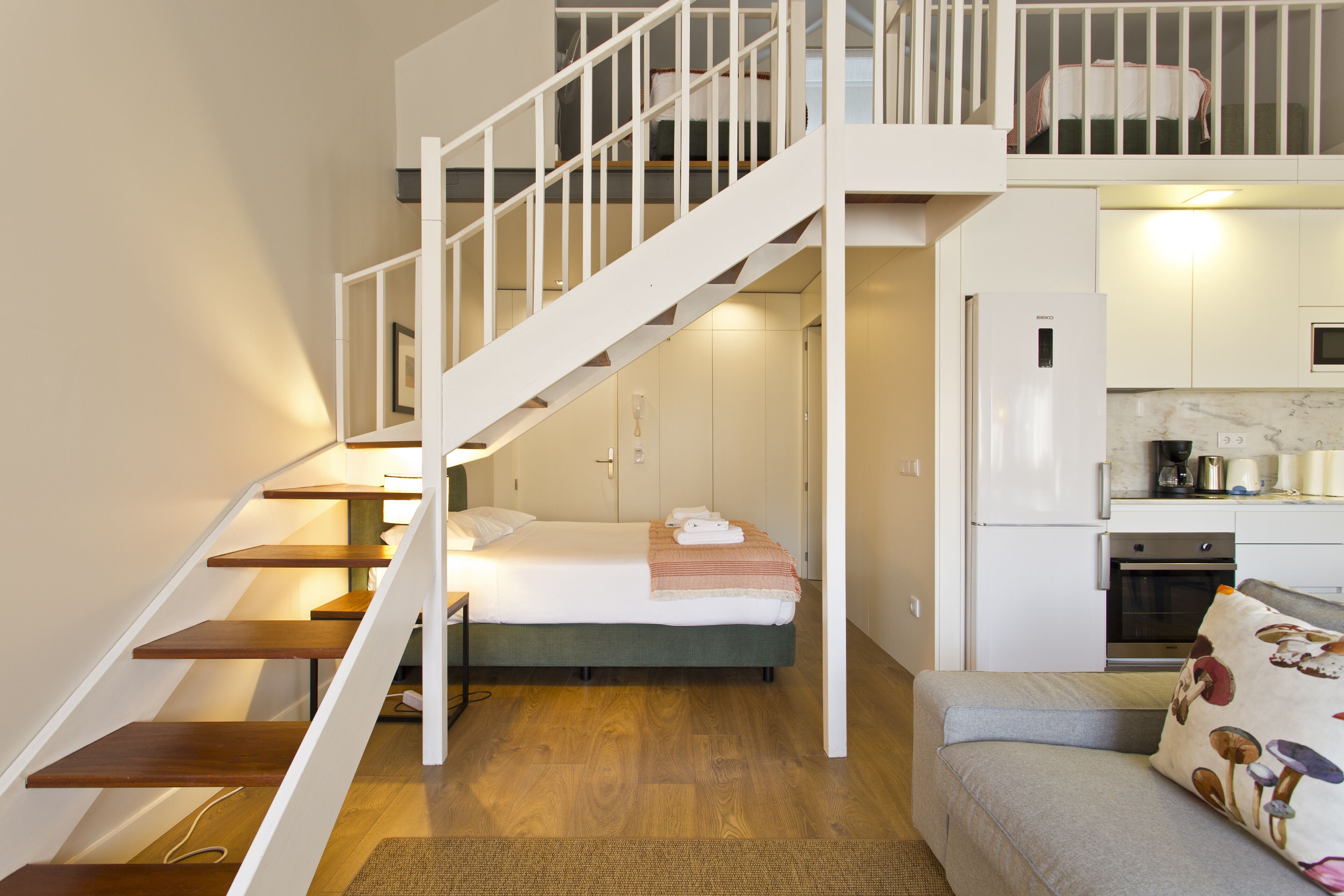 Ein kompakter Wohnraum mit Treppe zu einem Schlafzimmer im Zwischengeschoss, einer Küchenzeile und einer Sitzecke mit einer Couch.
