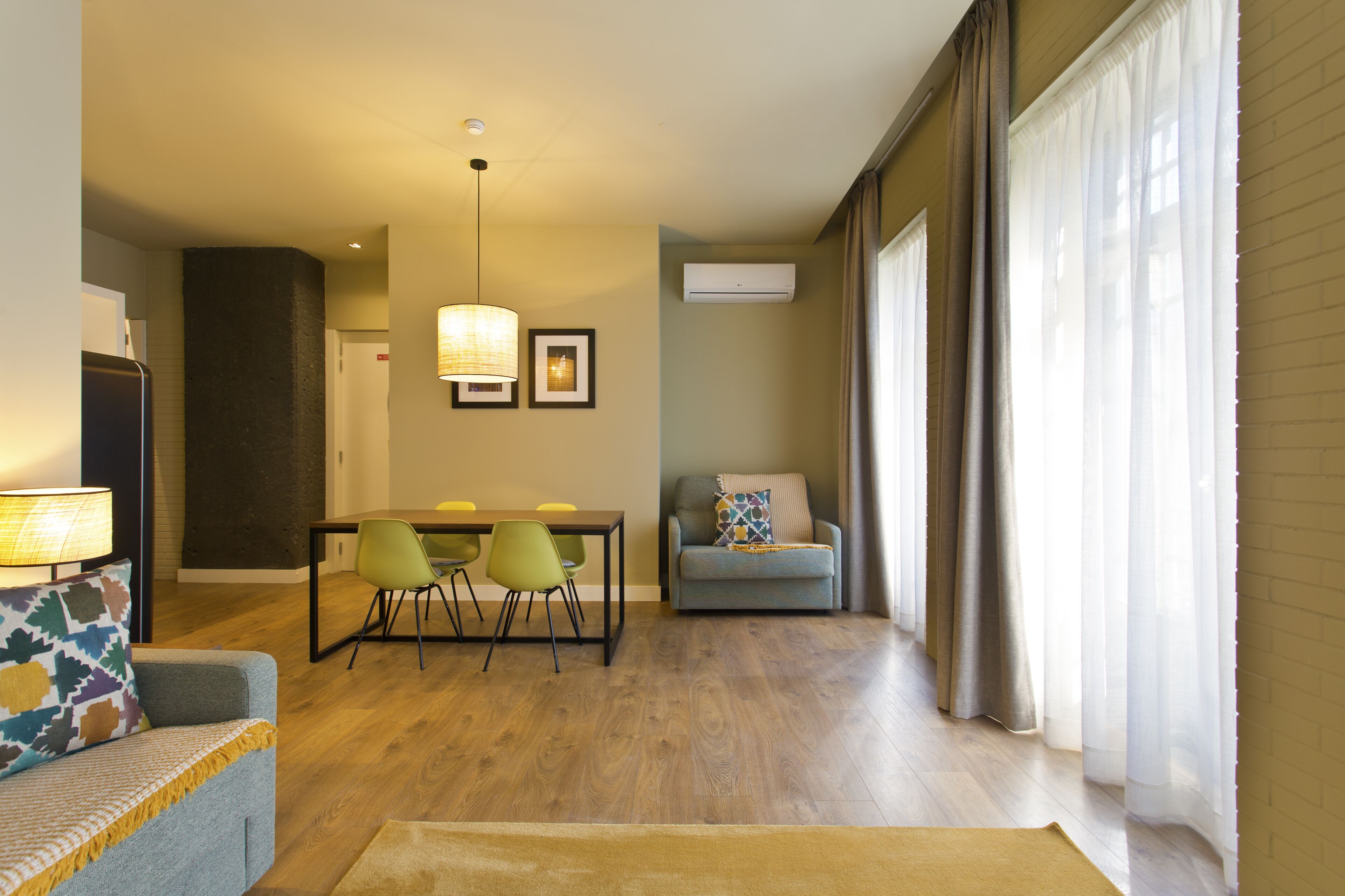 A modern living room with a sofa, a dining table with chairs, a lamp, and a large window with curtains.