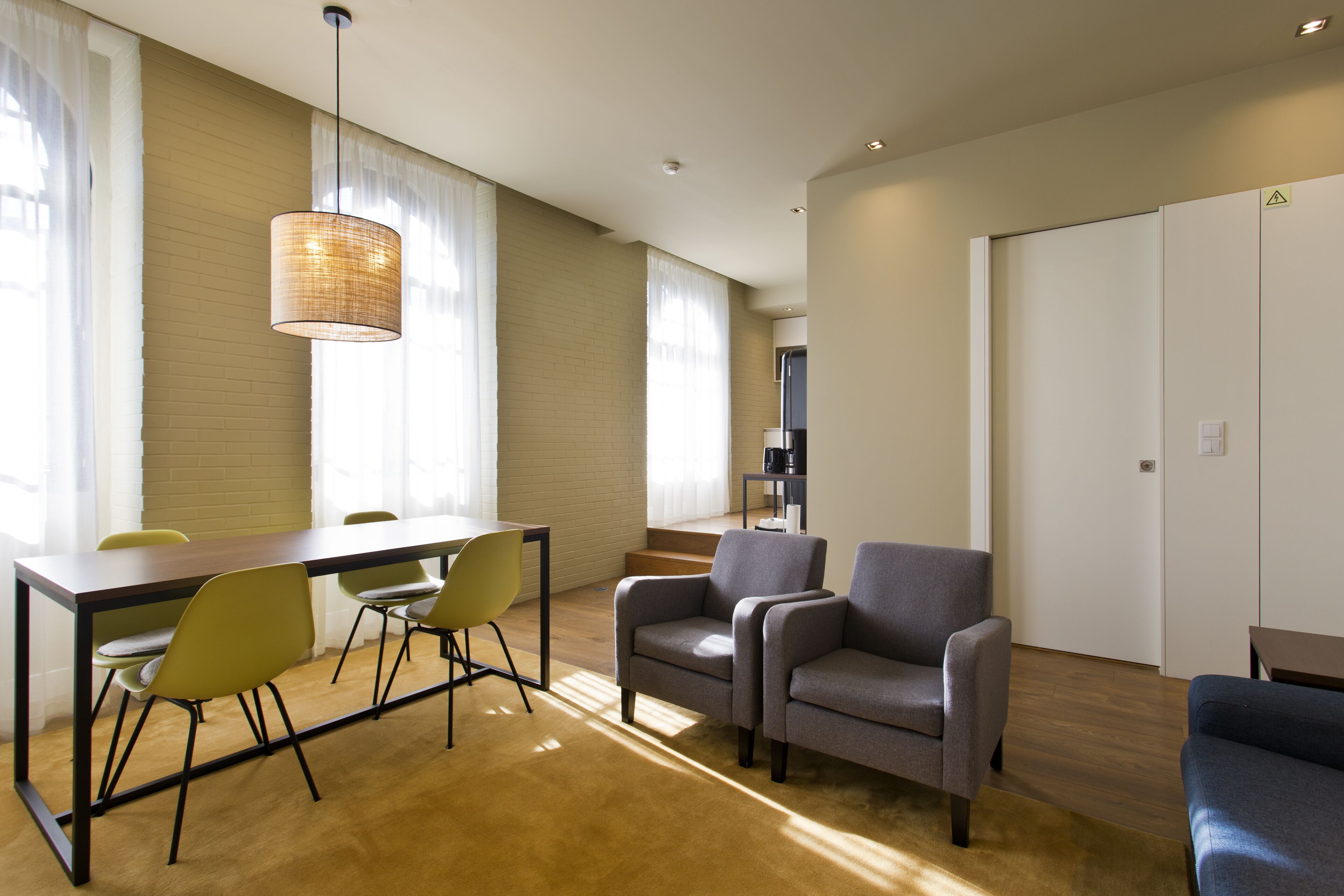 A modern living room with a dining area, featuring a wooden table, yellow chairs, grey armchairs, and a hanging light fixture.