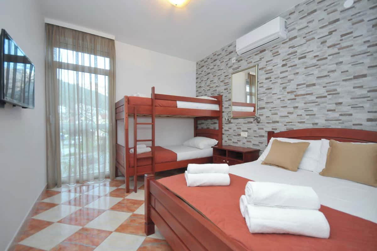 A hotel room with a bunk bed, a single bed, a wooden nightstand, a mirror, and a window with curtains.