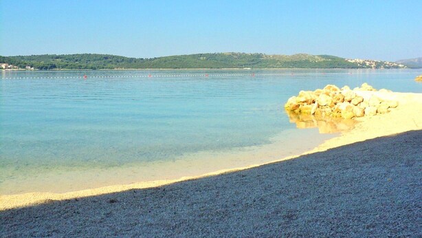 Vista despejada de una playa con agua tranquila, una orilla arenosa y una costa lejana con edificios.
