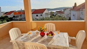 Un balcón con una mesa dispuesta para dos personas, que incluye un jarrón de flores, un cuenco de fruta y dos vasos de zumo de naranja.