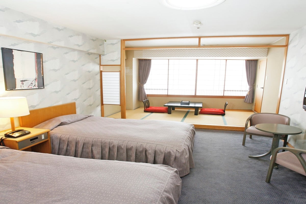 Quarto de hotel com duas camas, uma escrivaninha com abajur, uma mesinha, uma cadeira e uma televisão.