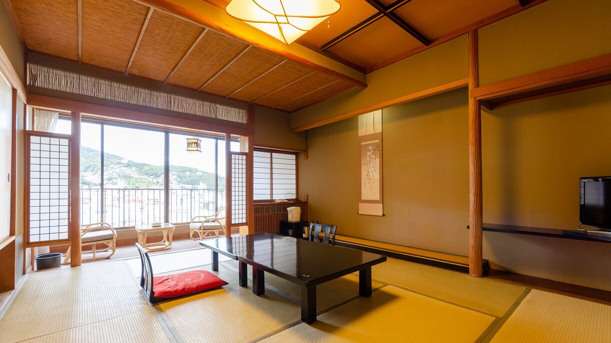 A tatami mat room with a low table, chairs, a television, and a view of a hillside.