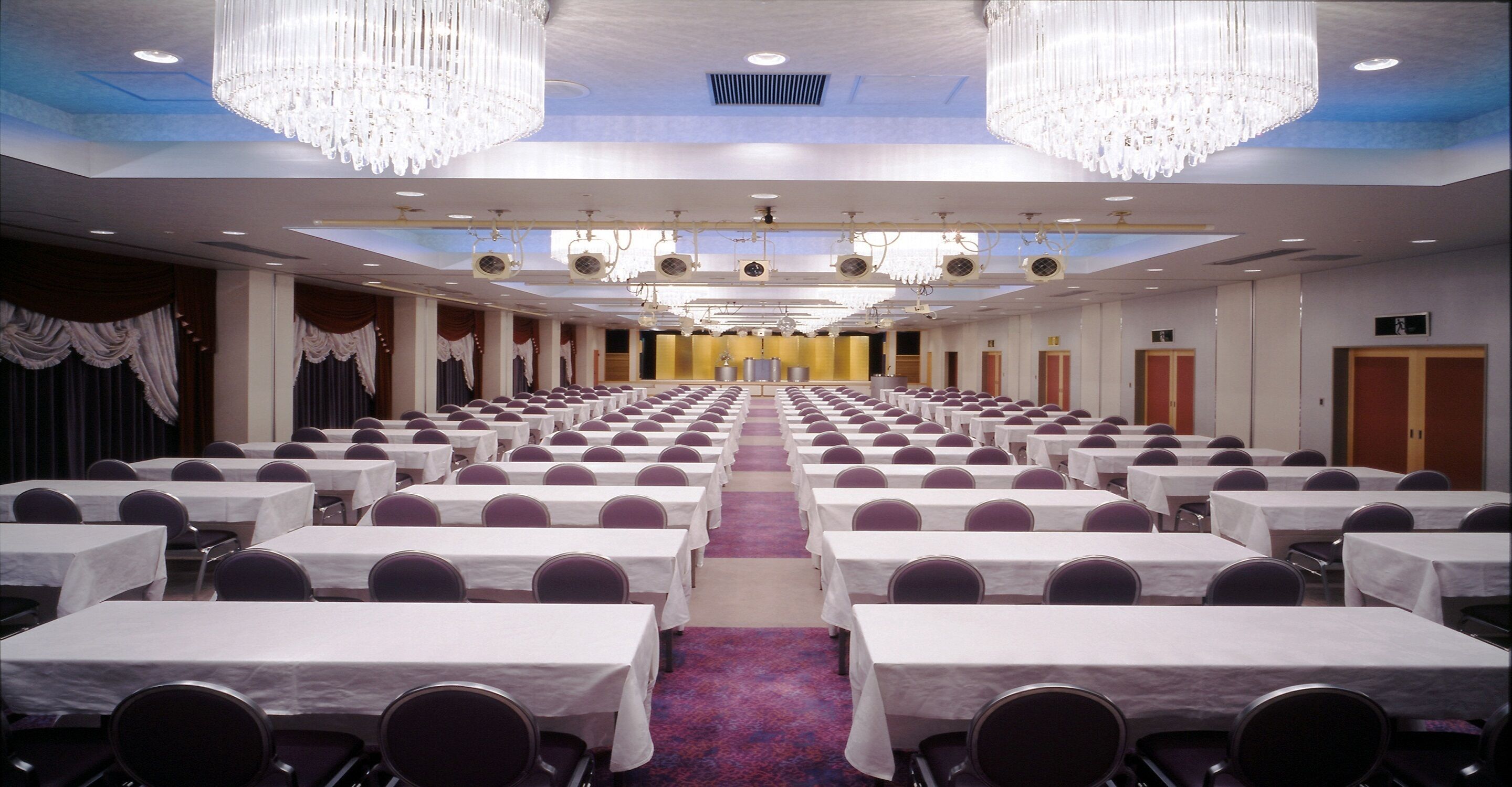 Een congreszaal met rijen tafels en stoelen, kroonluchters en een blauw plafond.