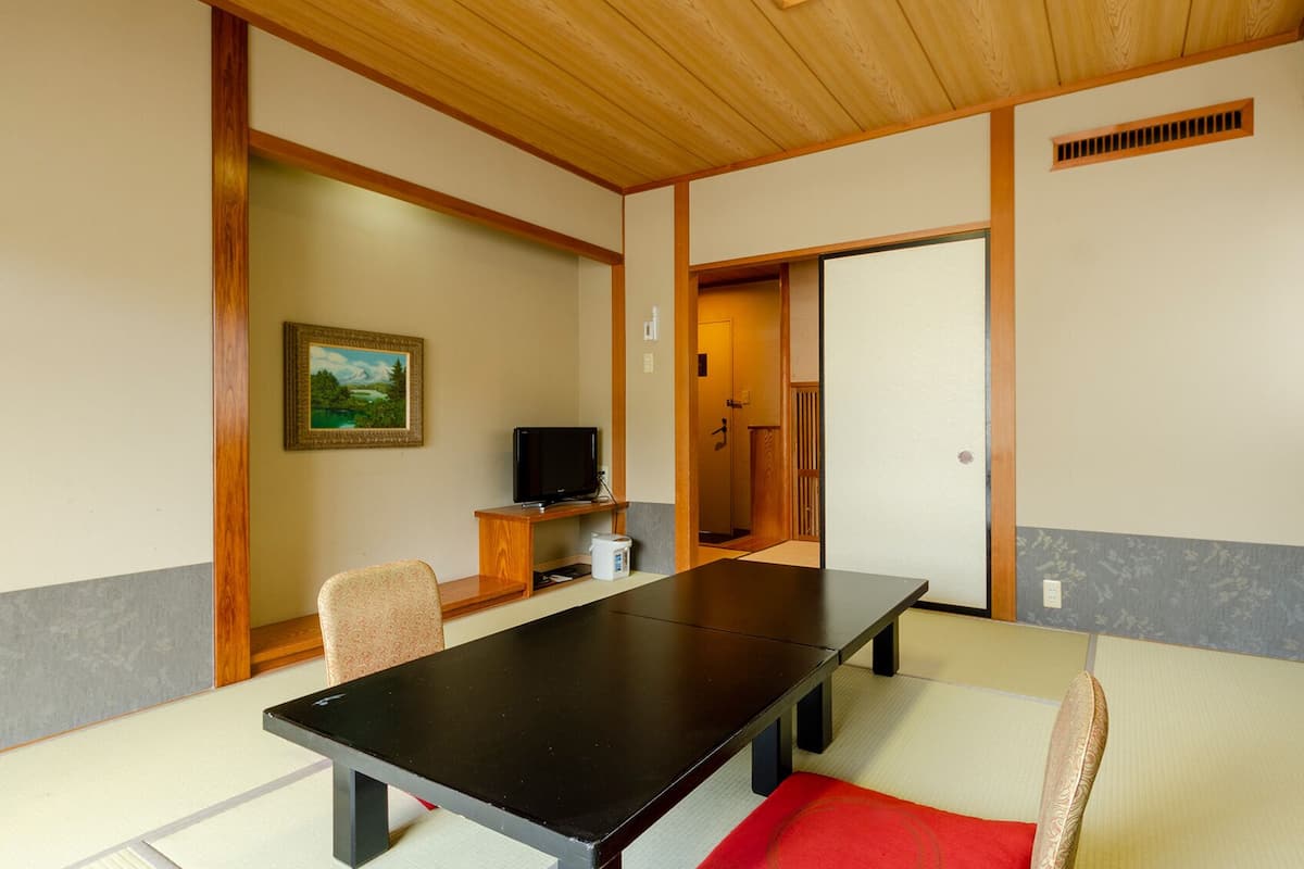 Um quarto tradicional de tatame com uma mesa baixa, cadeiras, uma televisão e um quadro na parede.