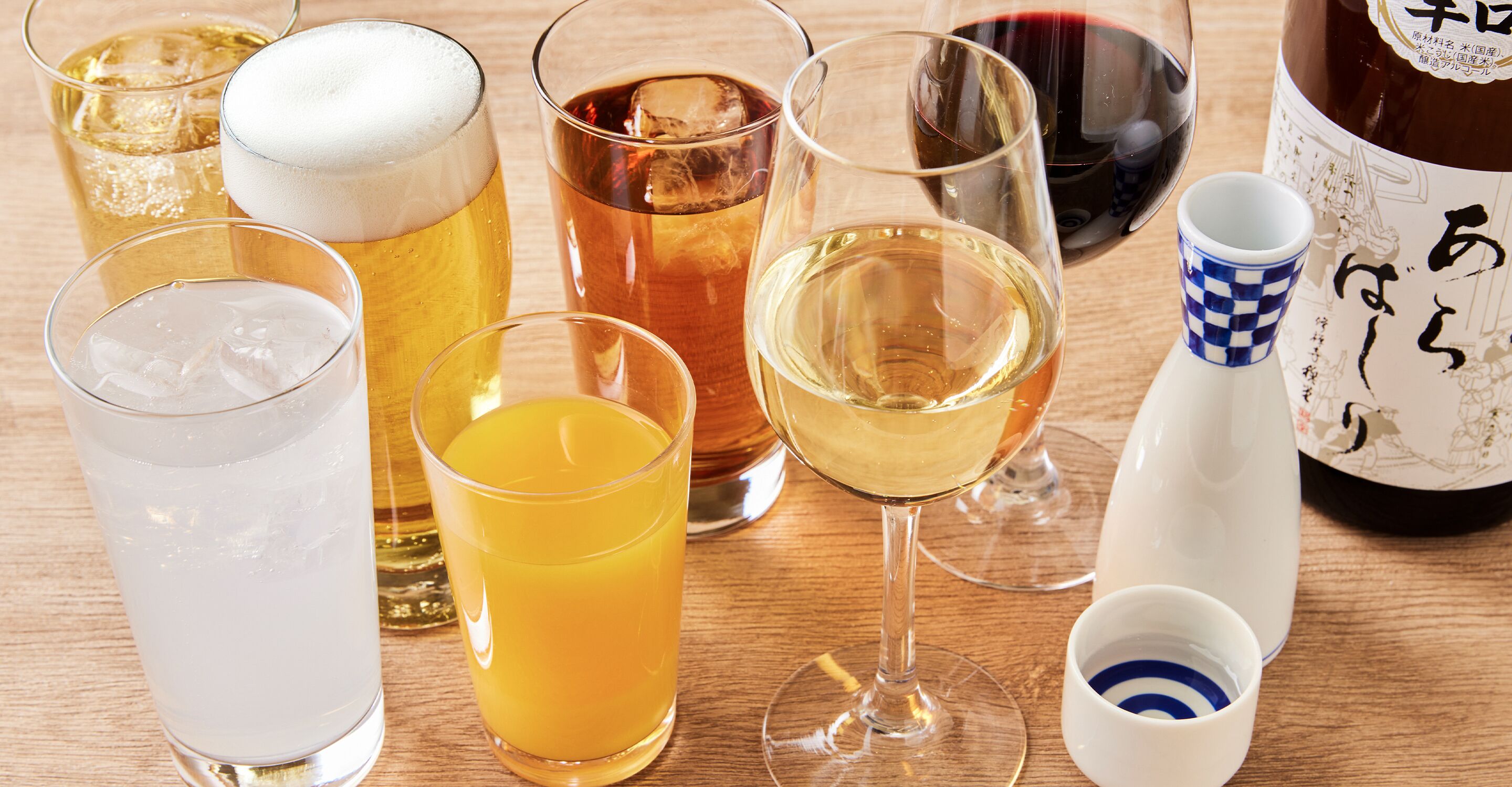 A variety of glasses containing different beverages, a bottle, and a teapot on a wooden surface.