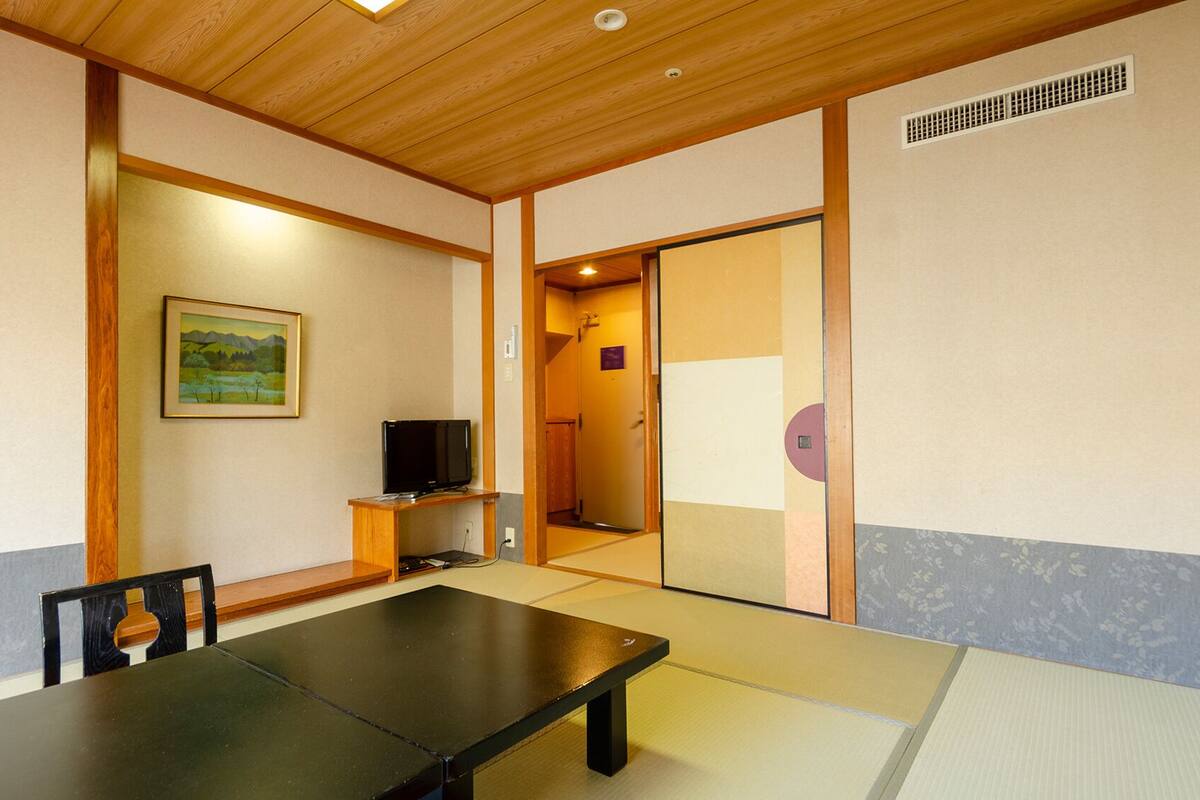 Um quarto japonês tradicional com piso de tatame, uma mesa baixa, uma televisão e uma porta deslizante para outro ambiente.