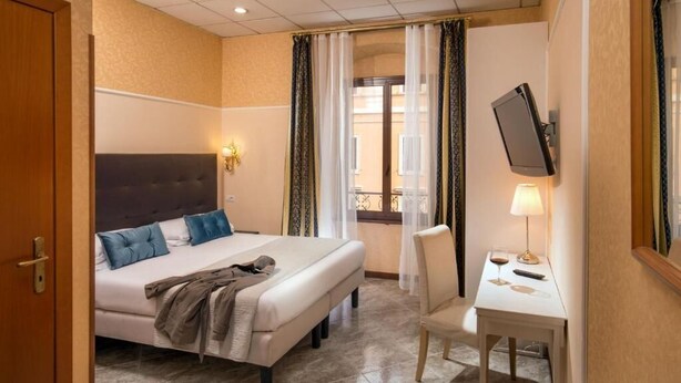 A hotel room with a bed, a desk with a lamp, a TV mounted on the wall, and a window with curtains. - Amalia Suites (Rome)